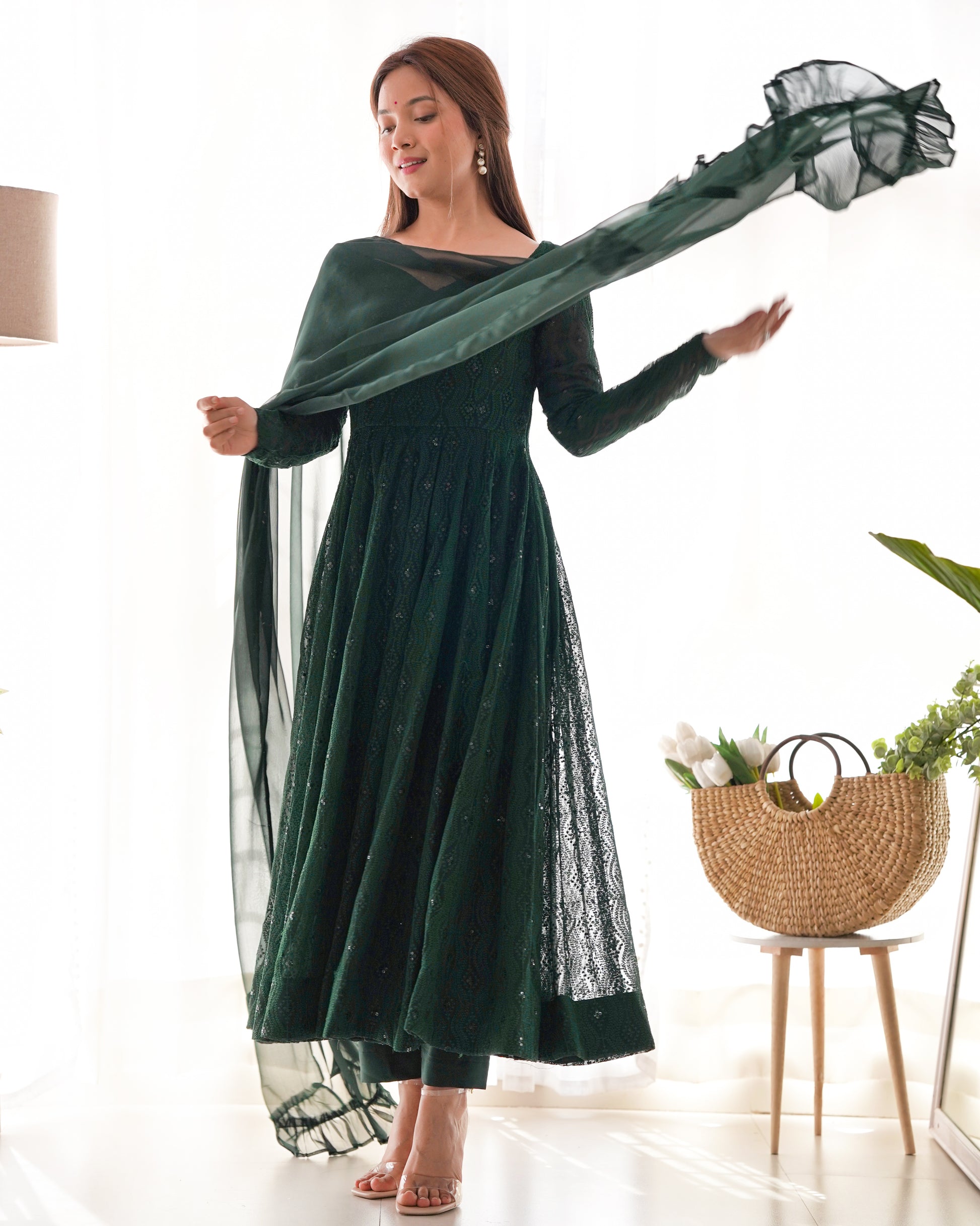 Woman in a green dress holding a sheer green scarf in a room with a basket and plant.