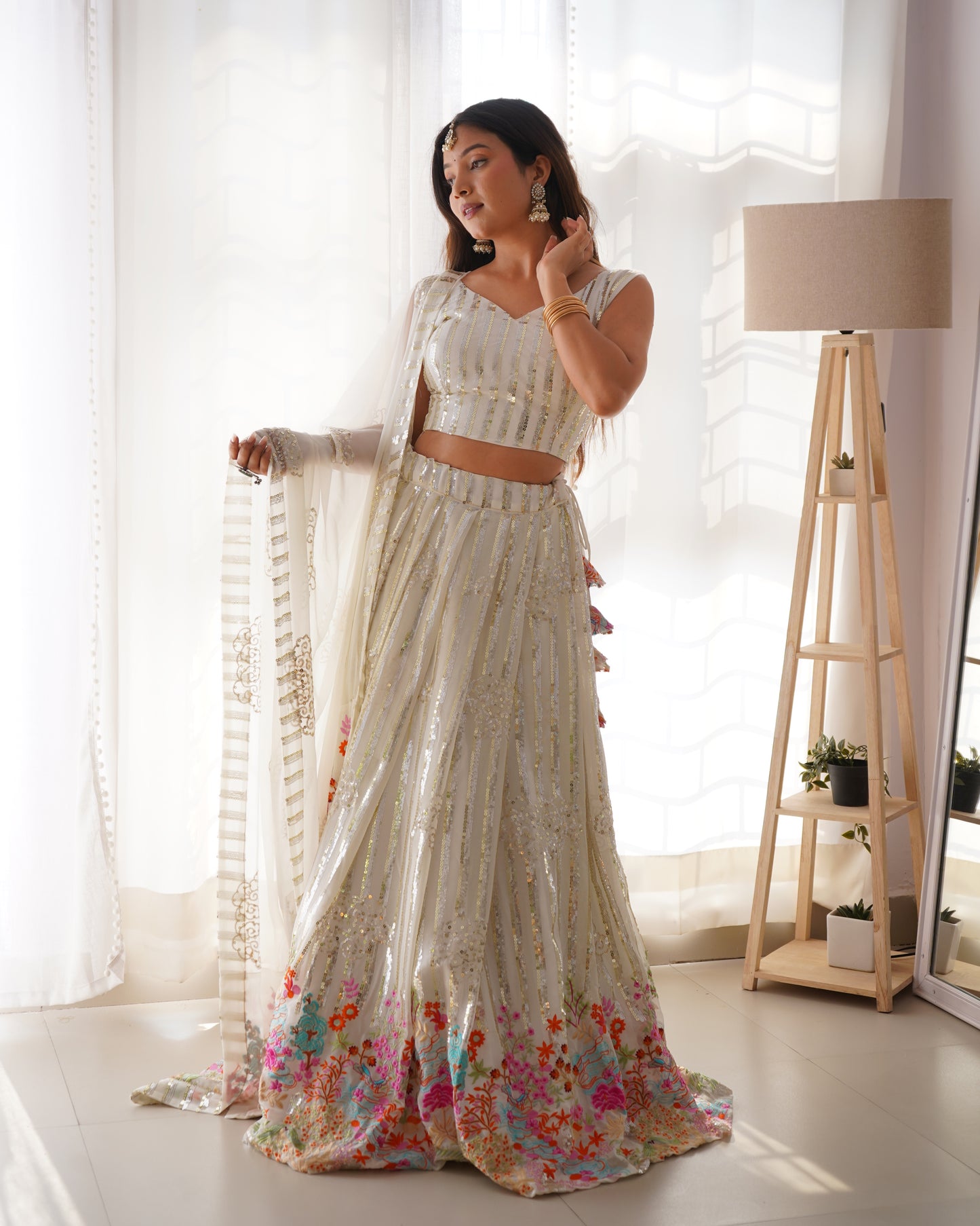 Woman in a floral saree standing in a bright room with white curtains and a lamp.