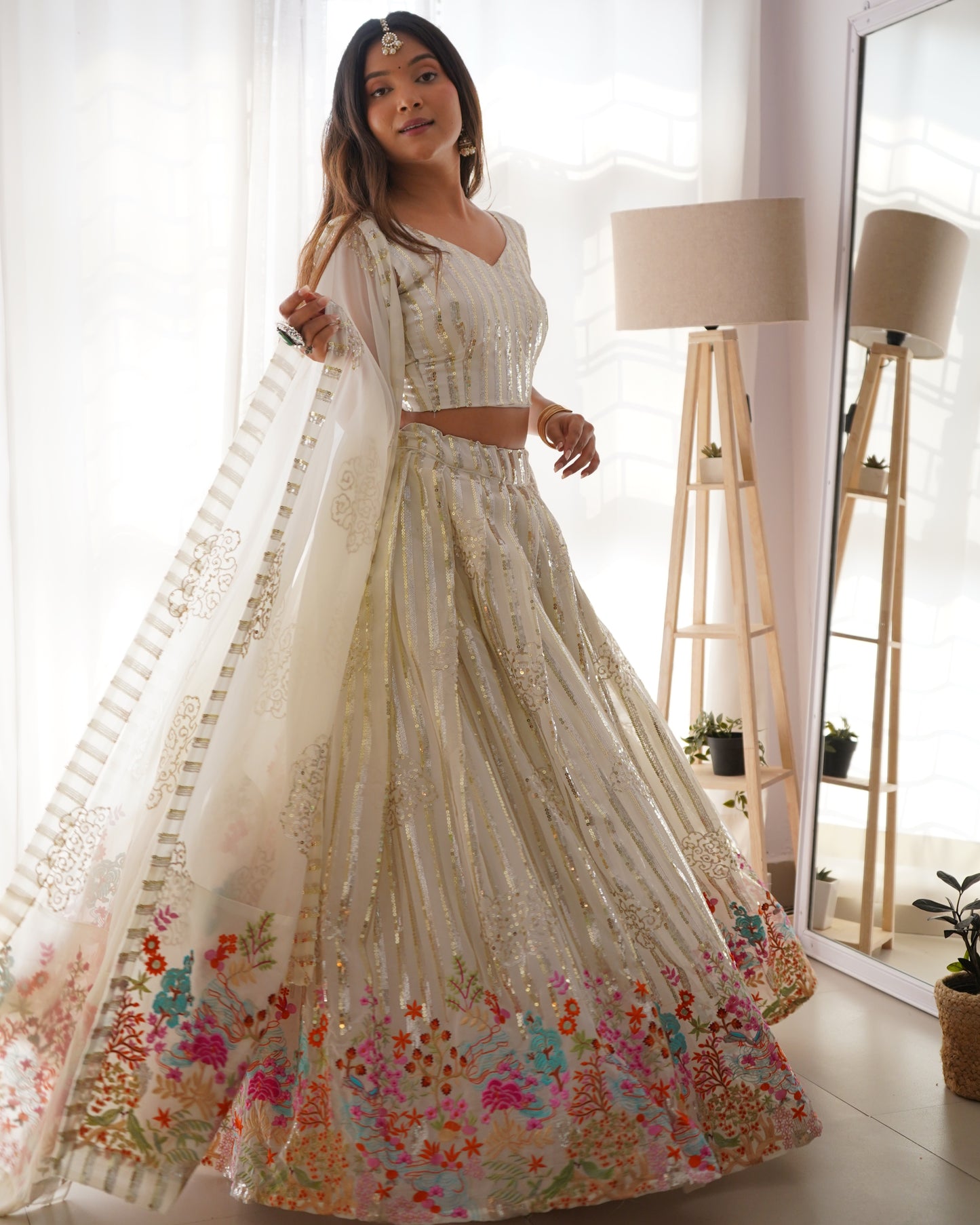 Woman in a white floral lehenga standing in a bright room with a mirror and lamp.