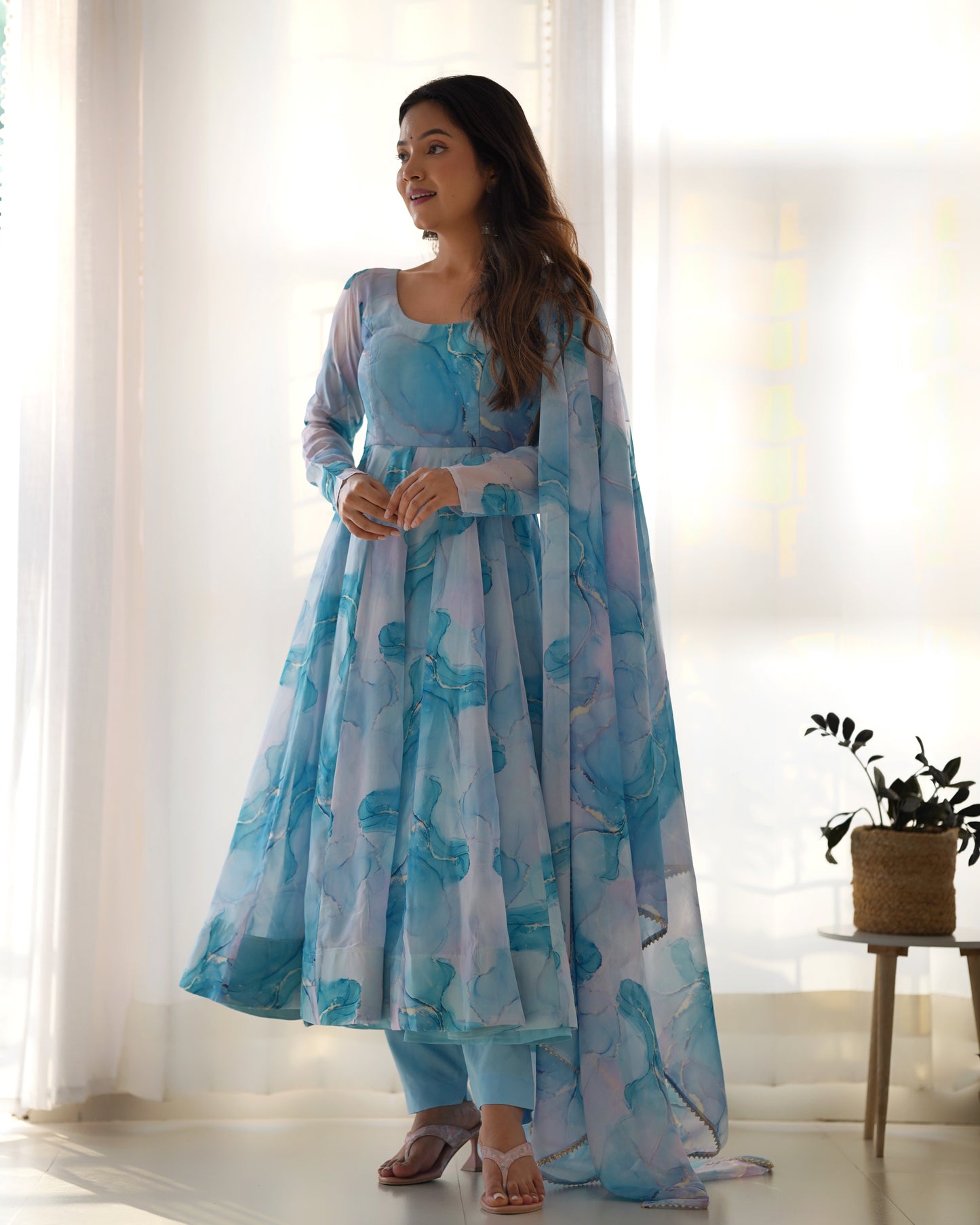 Woman wearing a light blue floral dress standing in a bright room with white curtains.