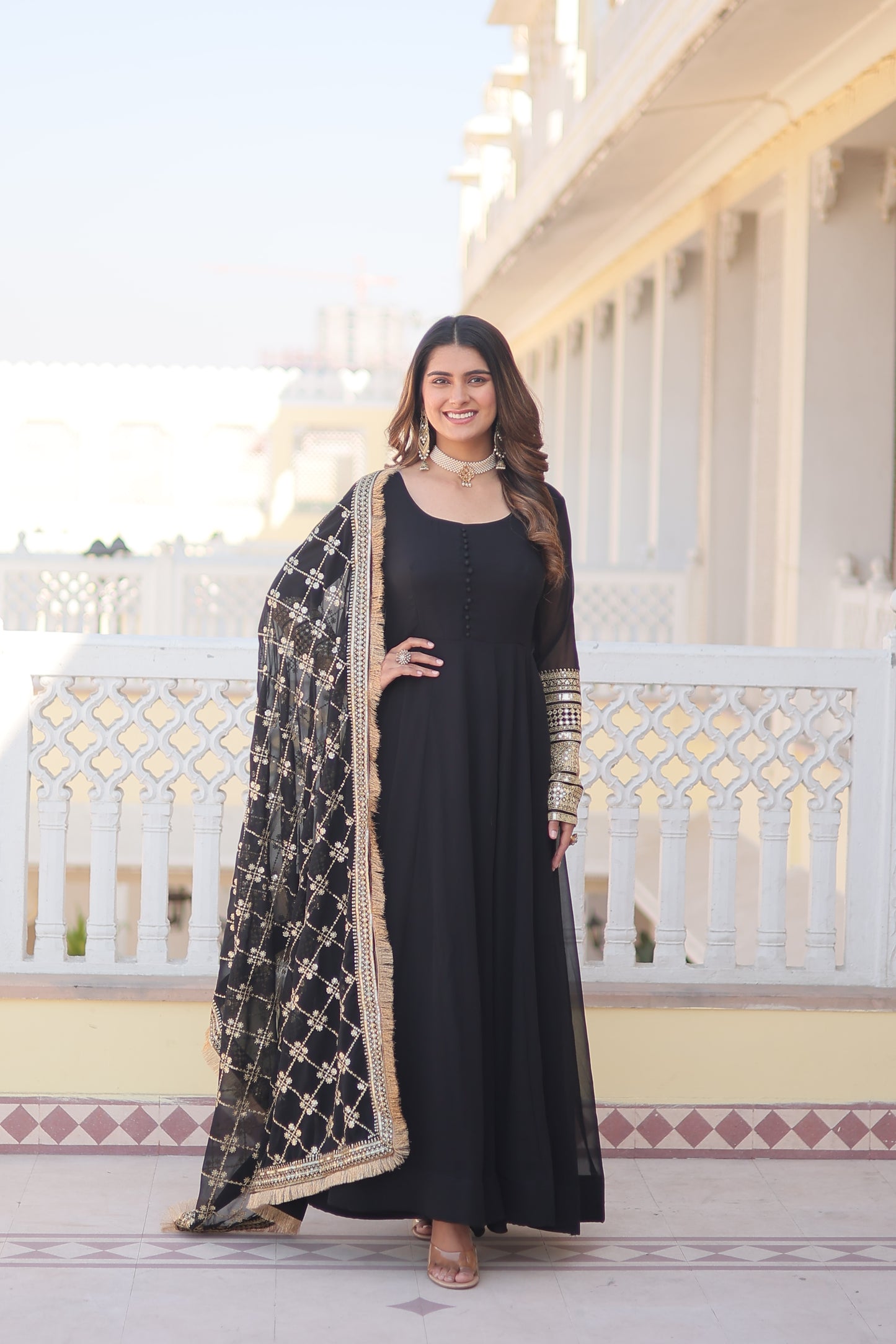 Woman in a black dress with intricate patterns standing in front of a decorative wall.