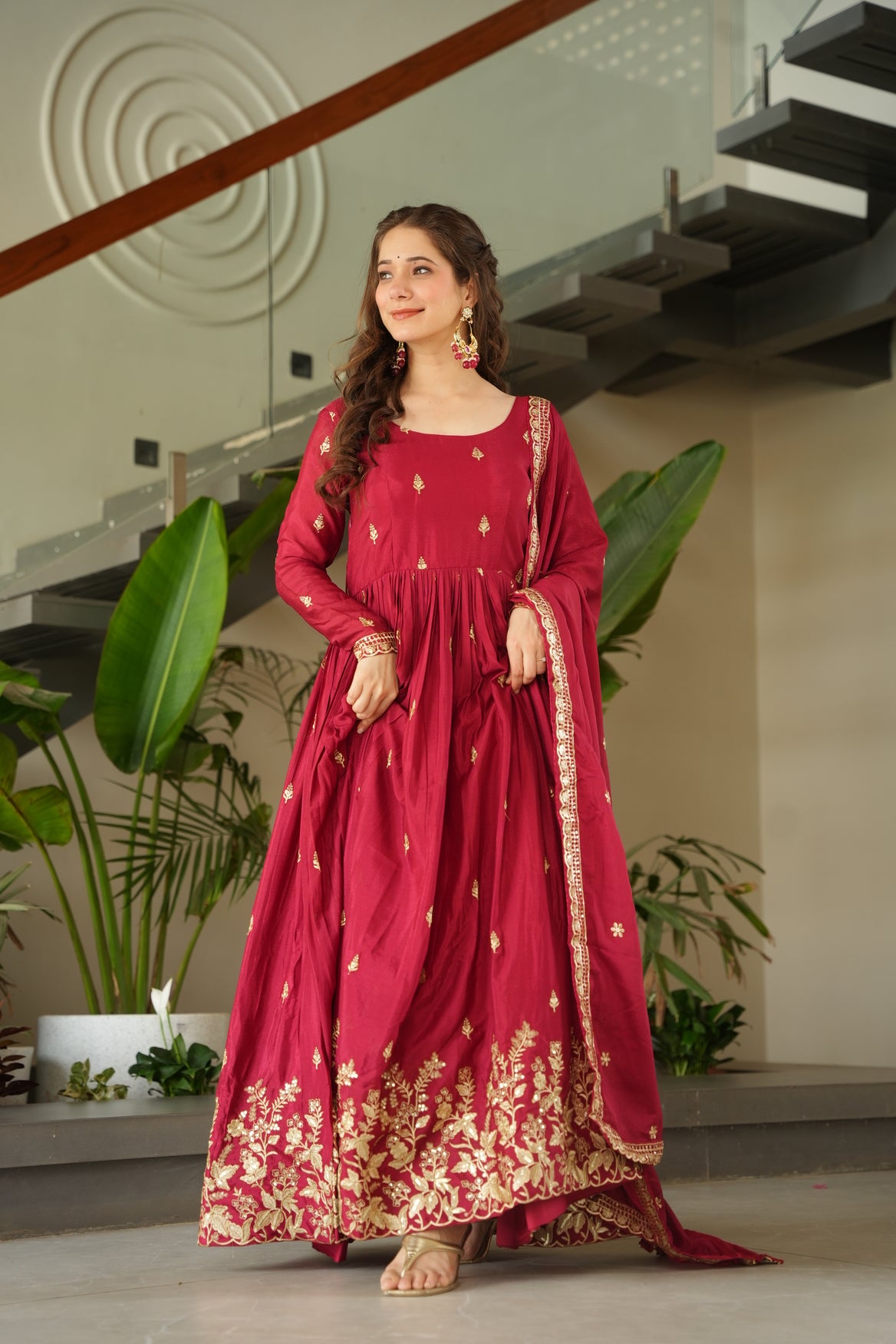 Woman in a red traditional outfit with gold patterns standing indoors.