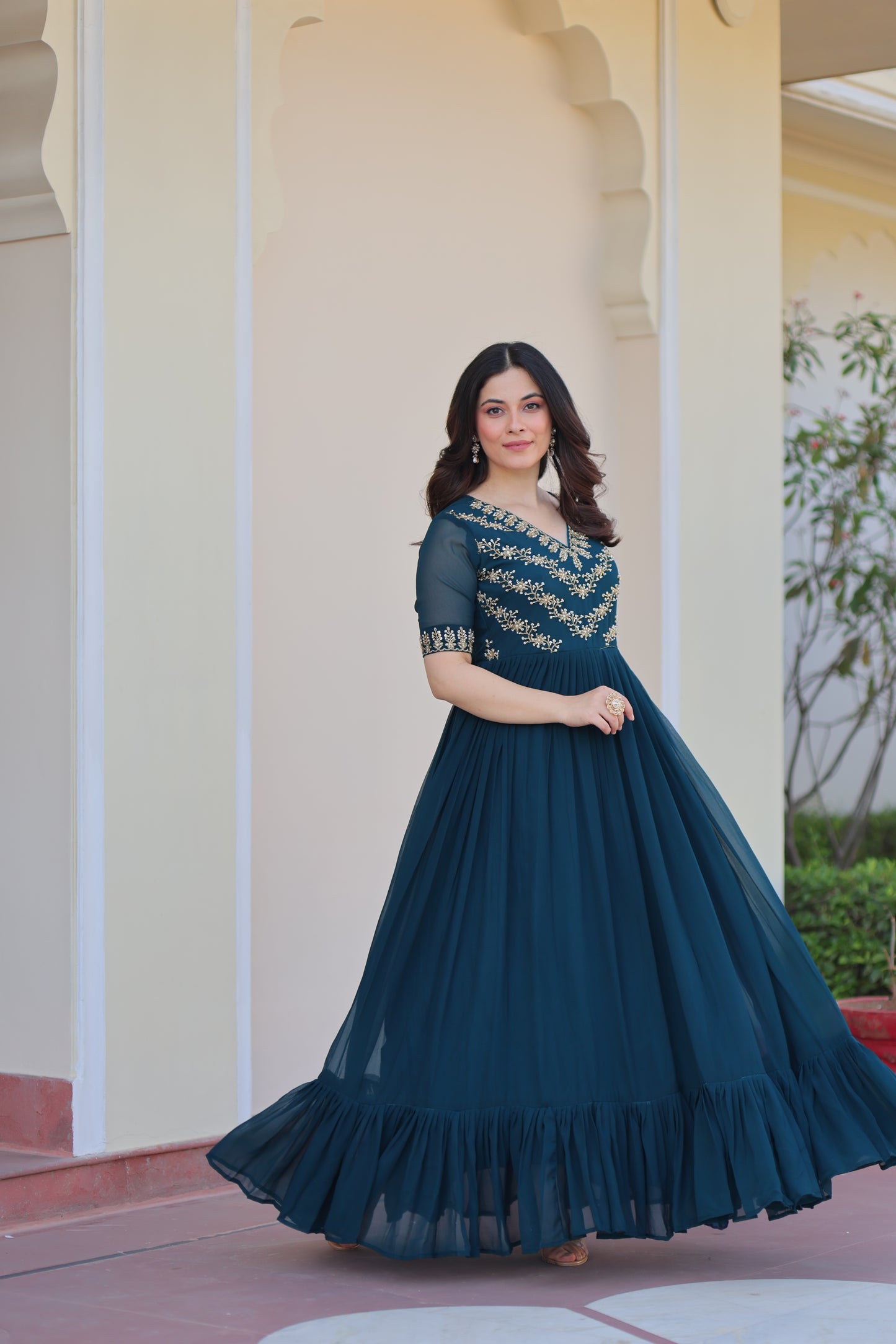 Woman in a long teal dress standing against a white wall.