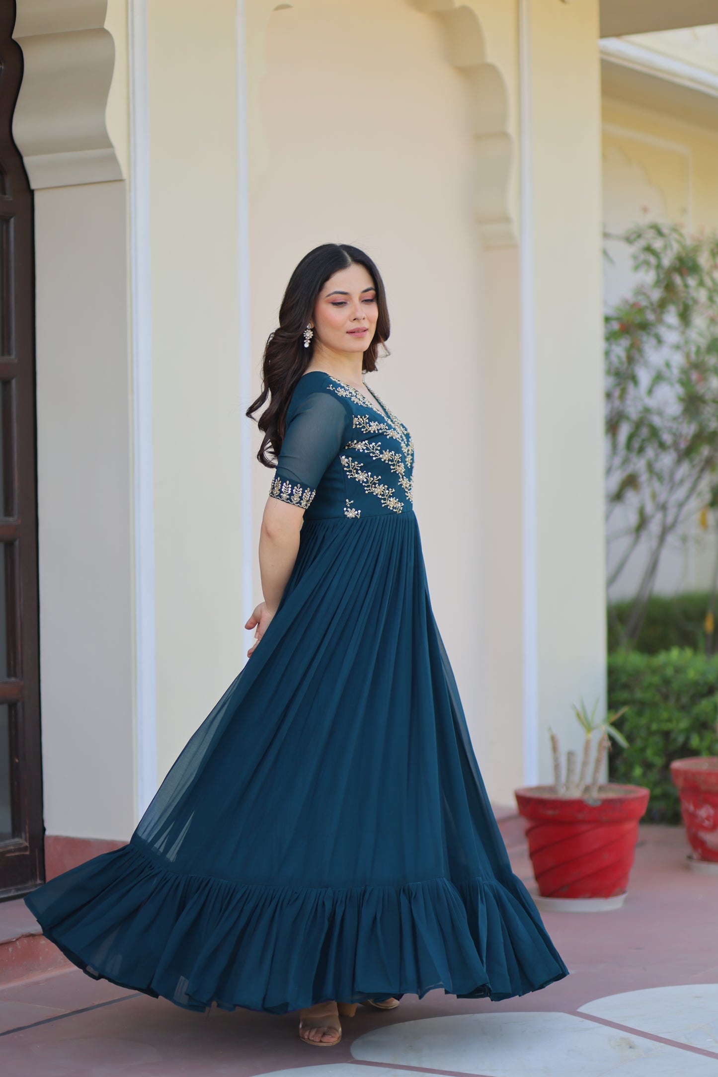 Woman wearing a blue evening gown with a pleated skirt, standing against a white wall.
