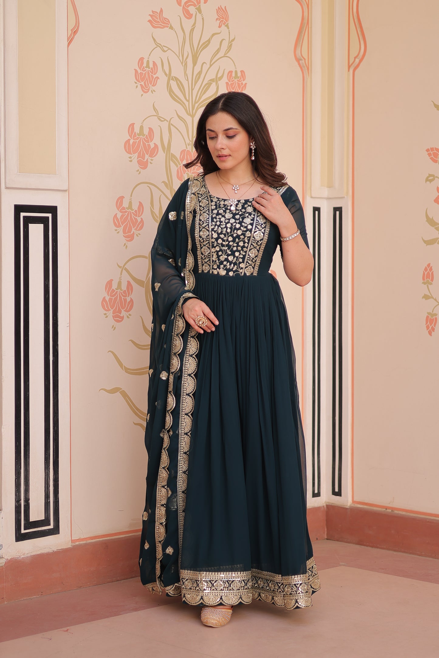 Woman in a dark blue dress with intricate patterns standing against a decorative wall.