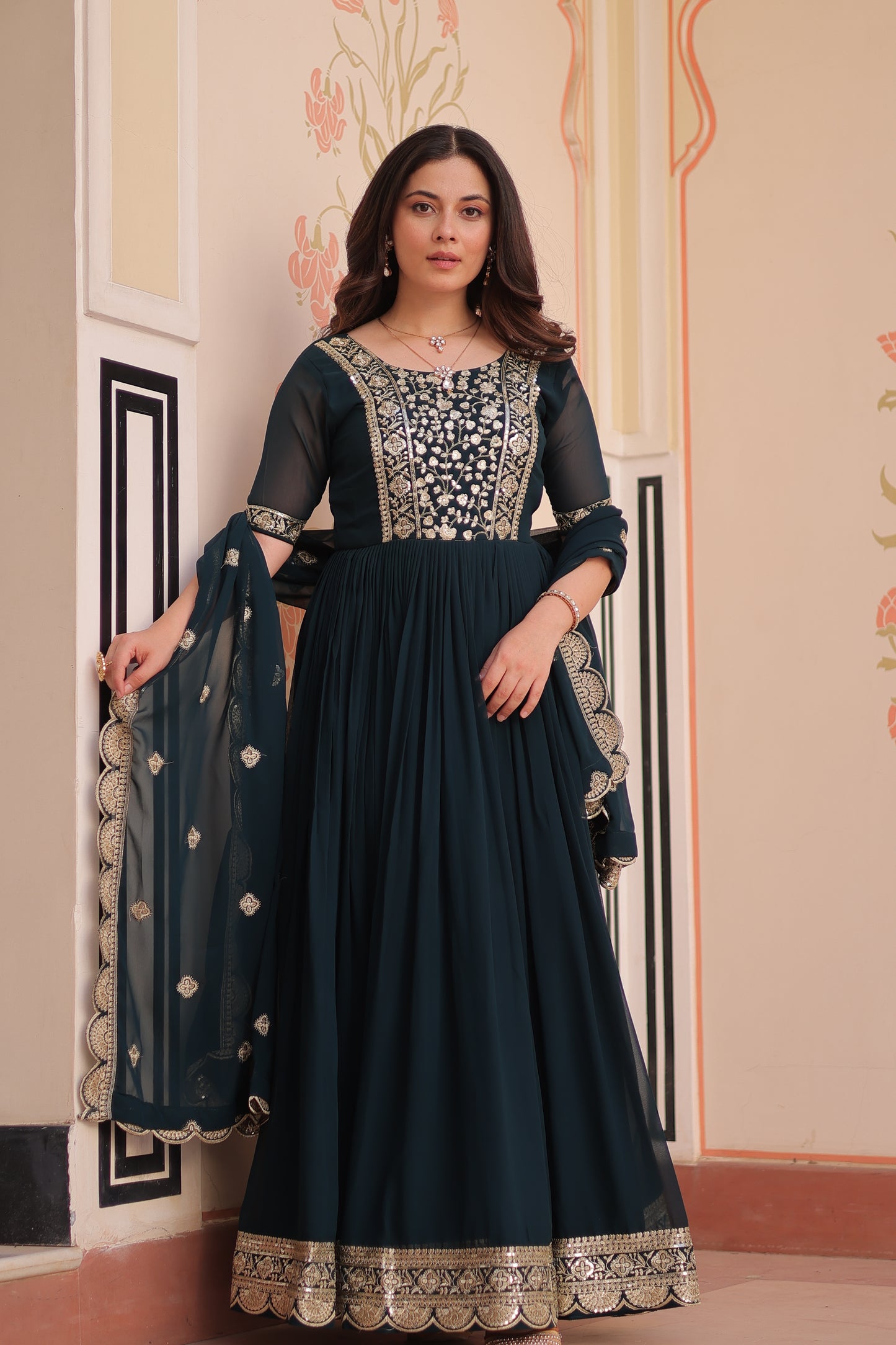 Woman wearing a dark blue dress with intricate patterns, standing against a decorative wall.