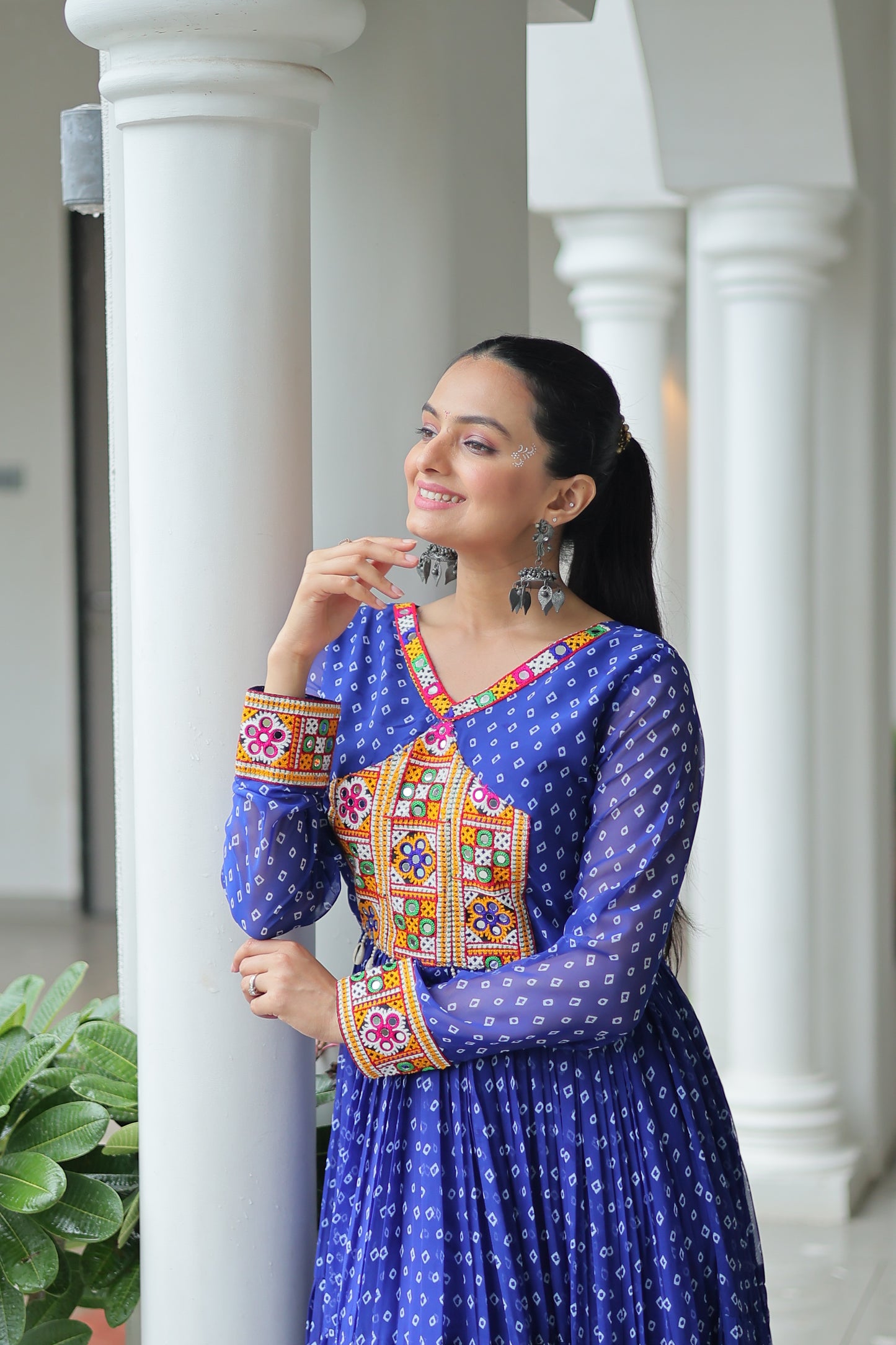Woman in traditional blue outfit with colorful patterns leaning against a white column.