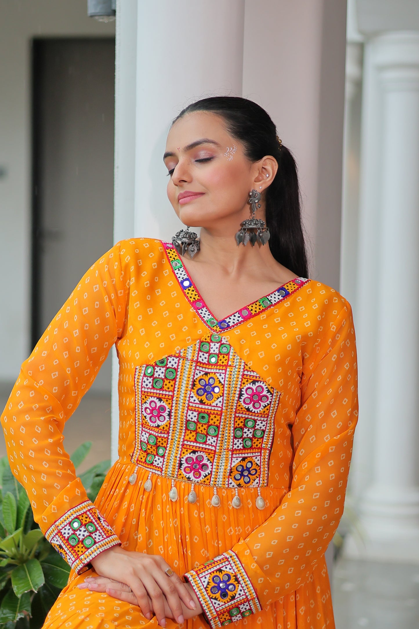 Woman wearing an orange traditional outfit with colorful embroidery, standing indoors.