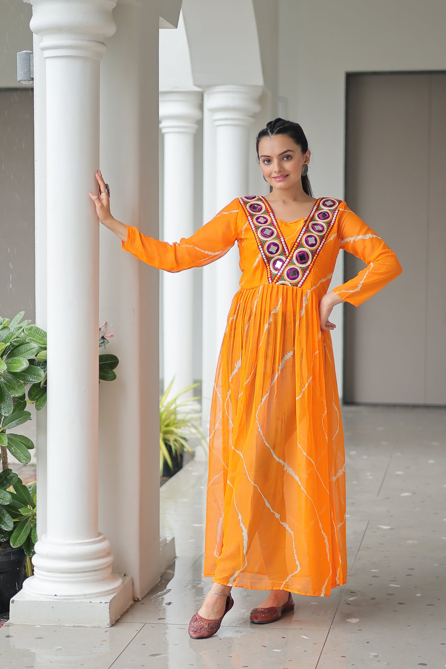 Woman in an orange traditional outfit standing indoors with white columns and greenery.