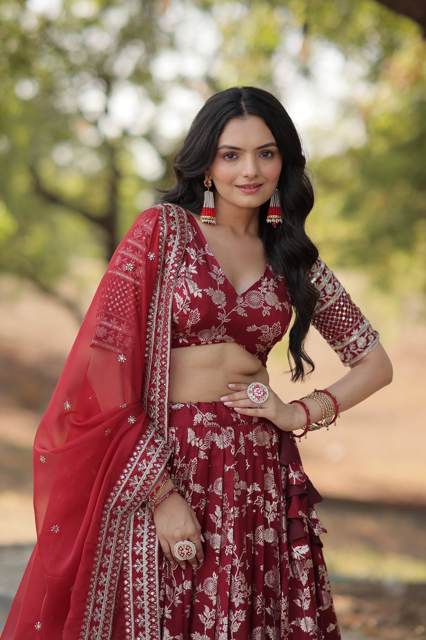 Woman in a red and white traditional outfit with floral patterns, standing outdoors with blurred greenery in the background.