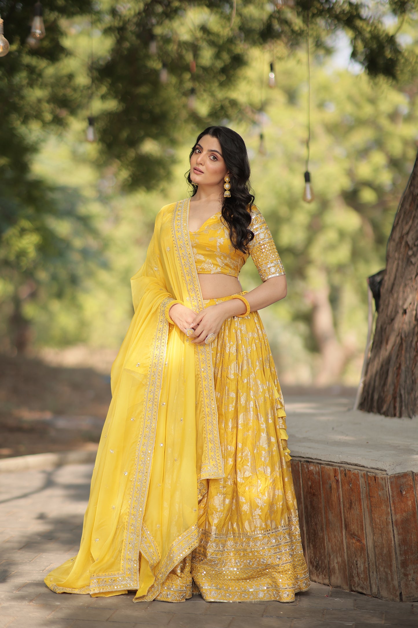 Woman in a yellow traditional outfit standing outdoors with greenery in the background