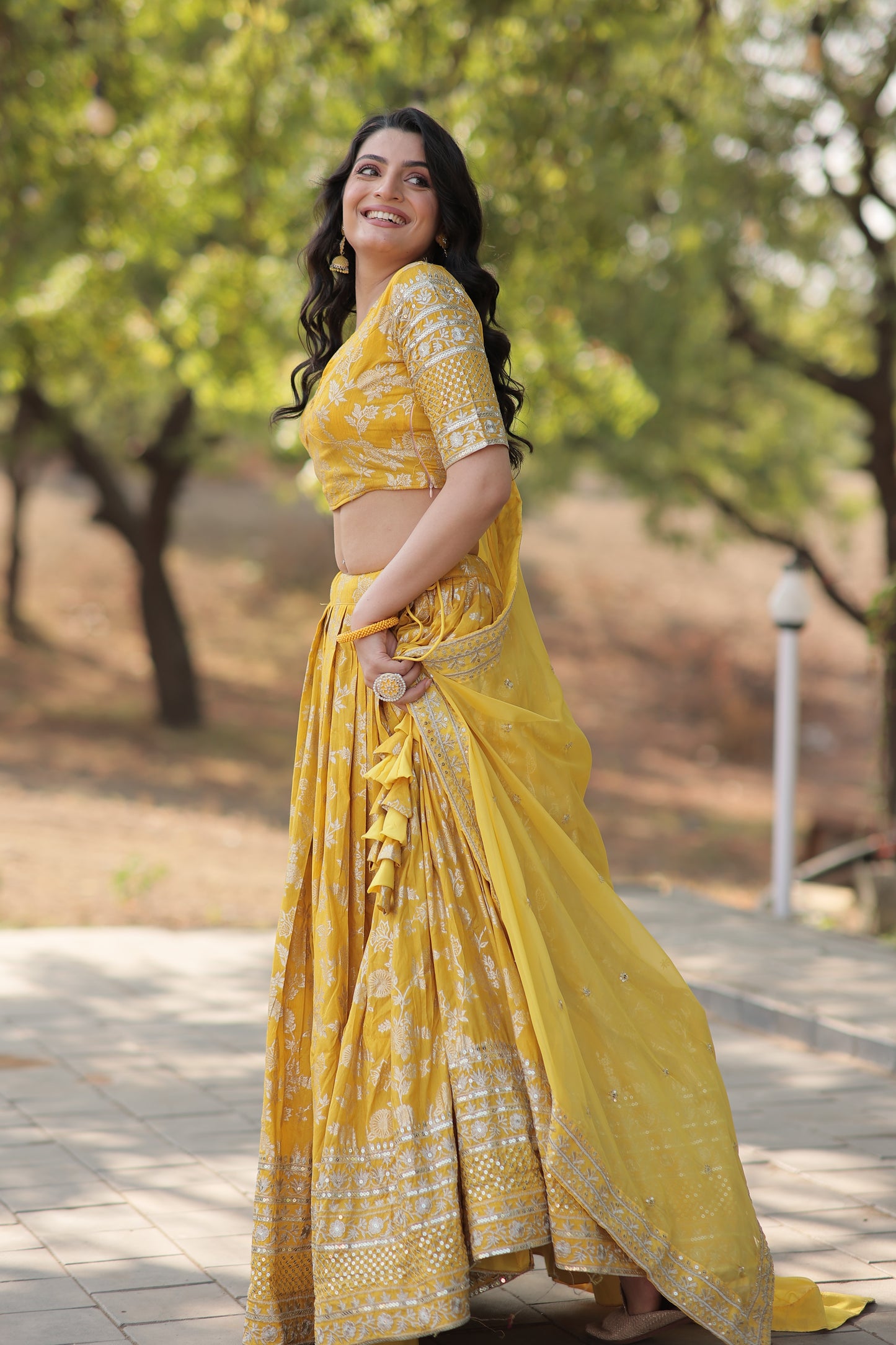 Woman in a yellow saree standing outdoors with trees in the background