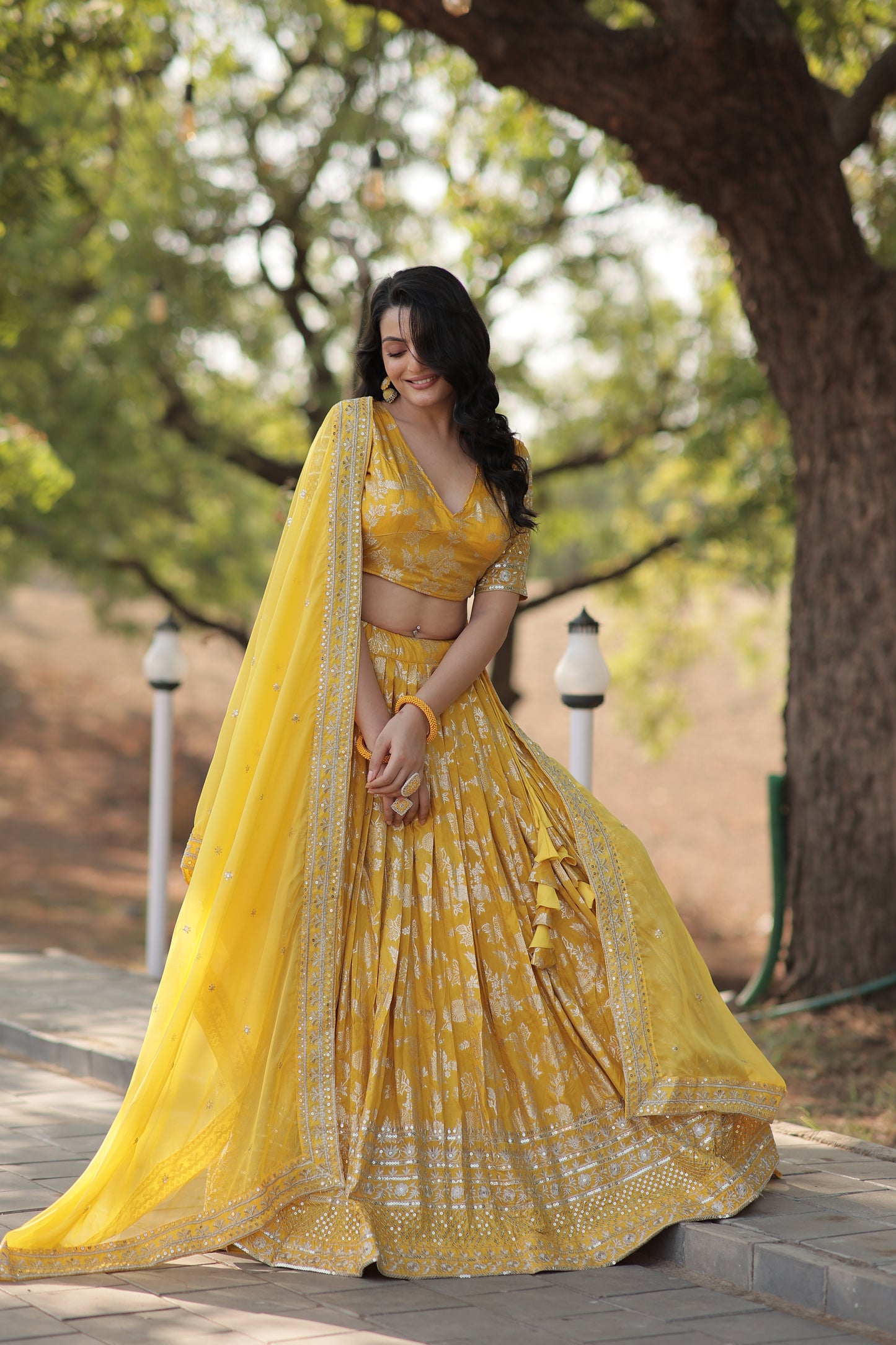 Woman in a yellow saree standing outdoors with trees in the background