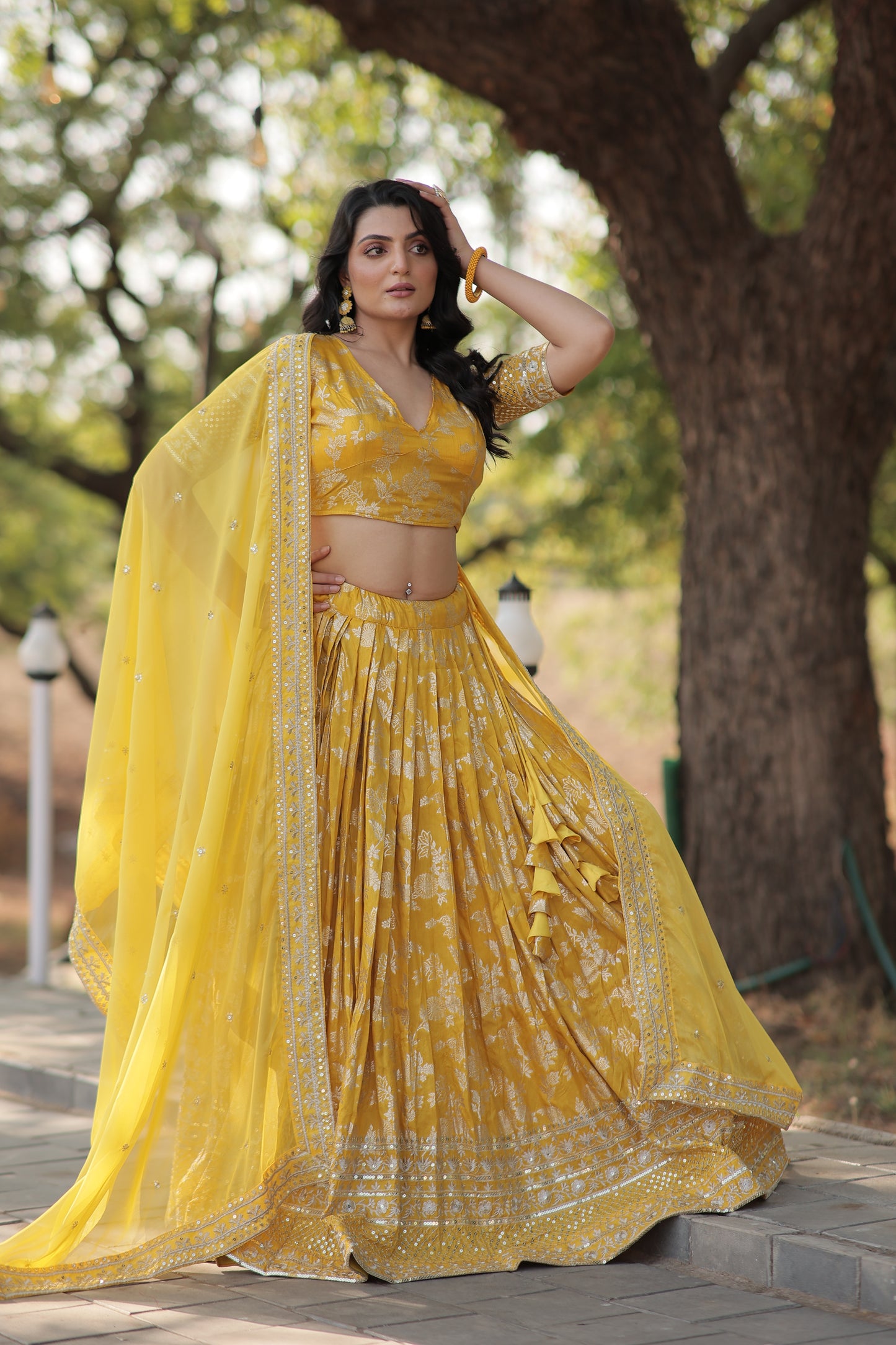 Woman in a yellow saree standing outdoors with trees in the background