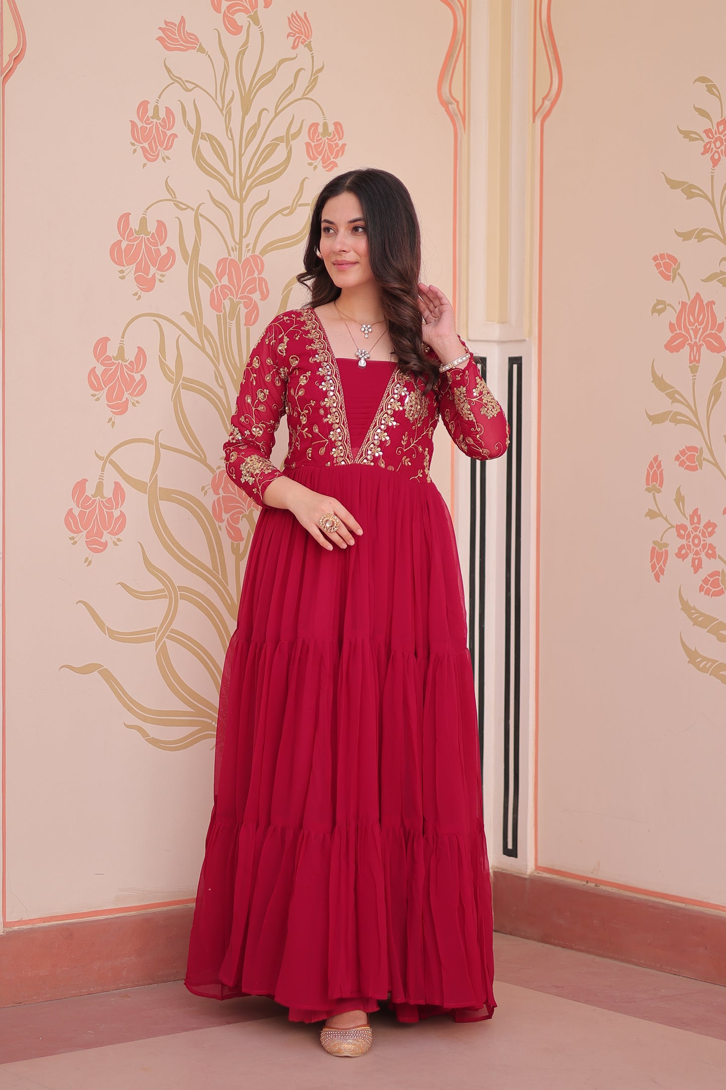 Woman in a red dress with floral patterns standing against a decorative wall.