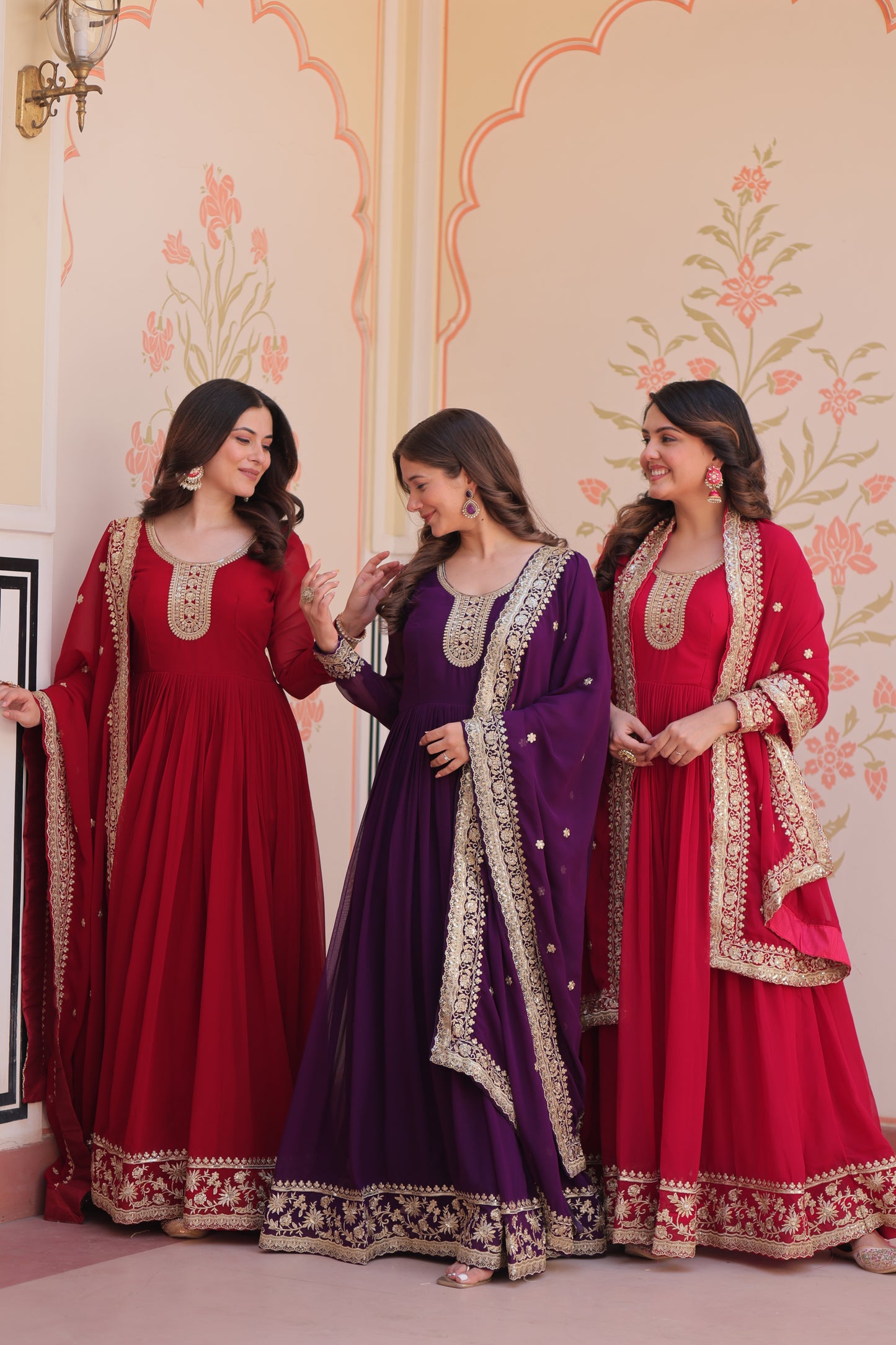 Three women in traditional red and purple dresses standing against a decorative wall.