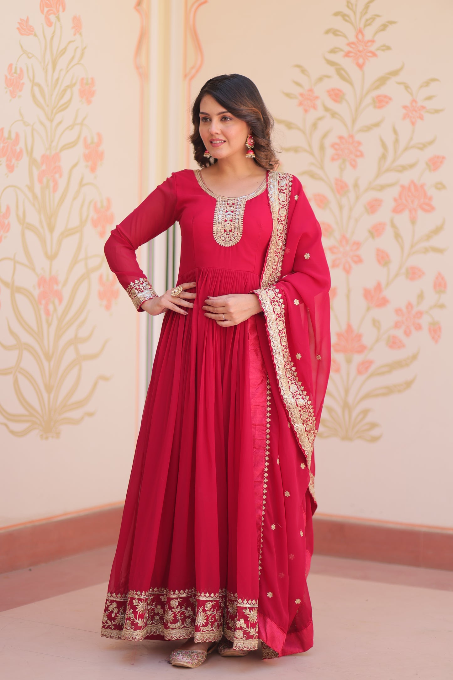 Woman wearing a red traditional outfit with gold embroidery on a floral patterned wall.