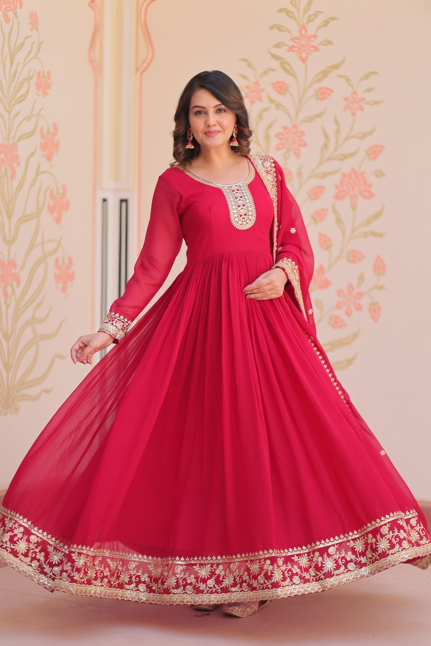 Woman in a red traditional outfit with gold embroidery standing against a floral wallpapered wall.