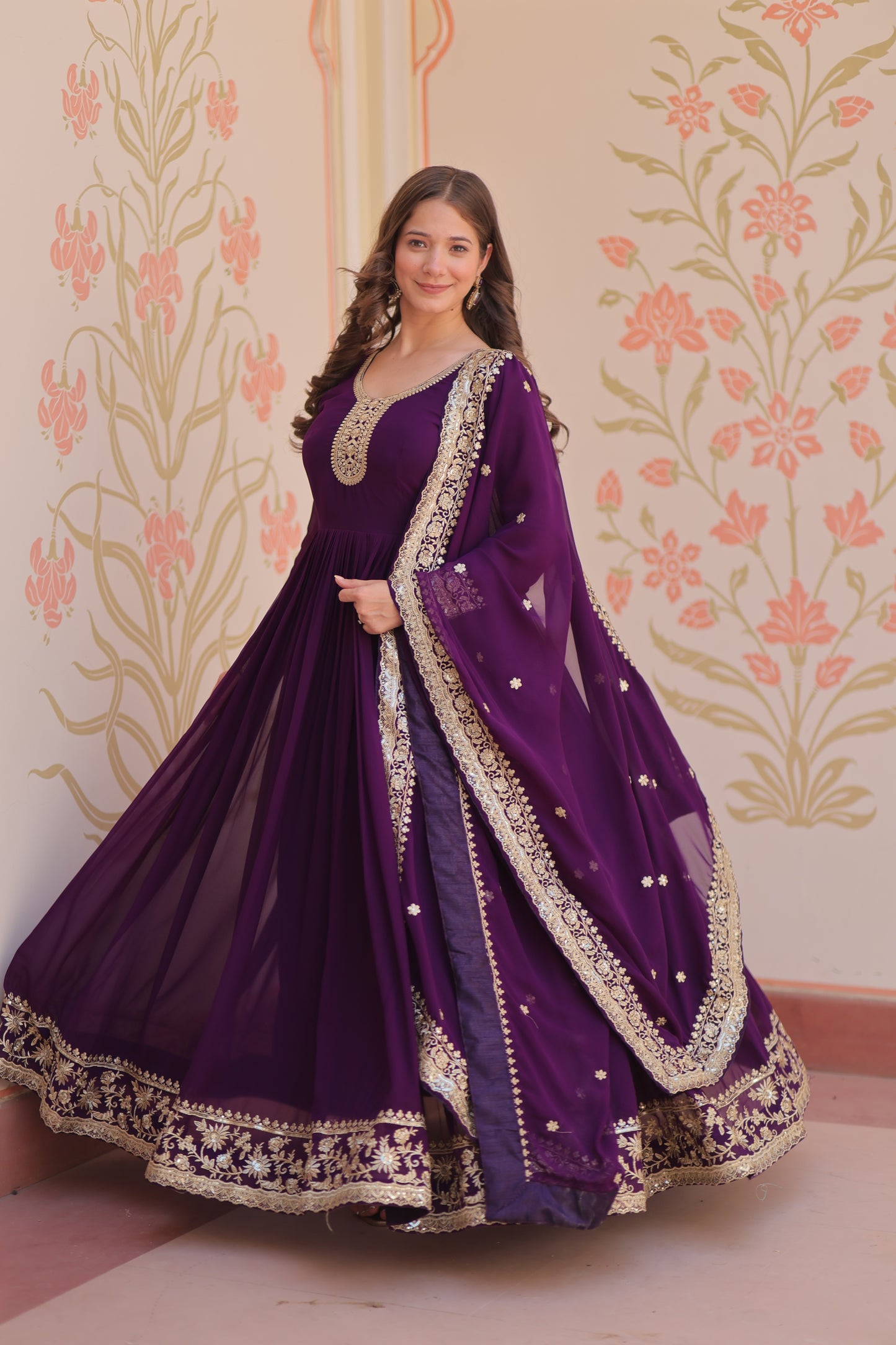 Woman wearing a purple dress with intricate detailing against a floral patterned wall.