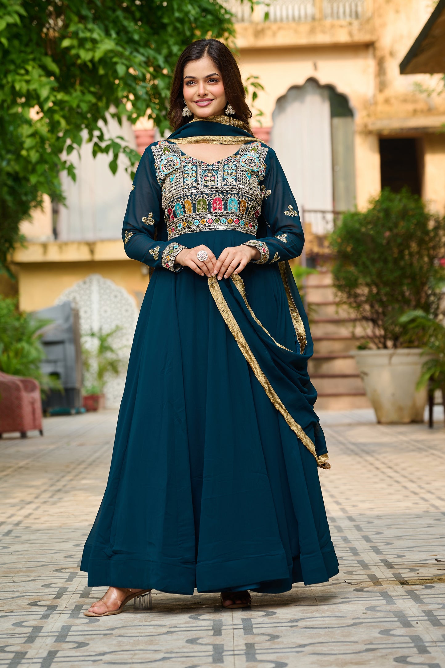 Woman in a traditional teal outfit with intricate patterns standing outdoors.