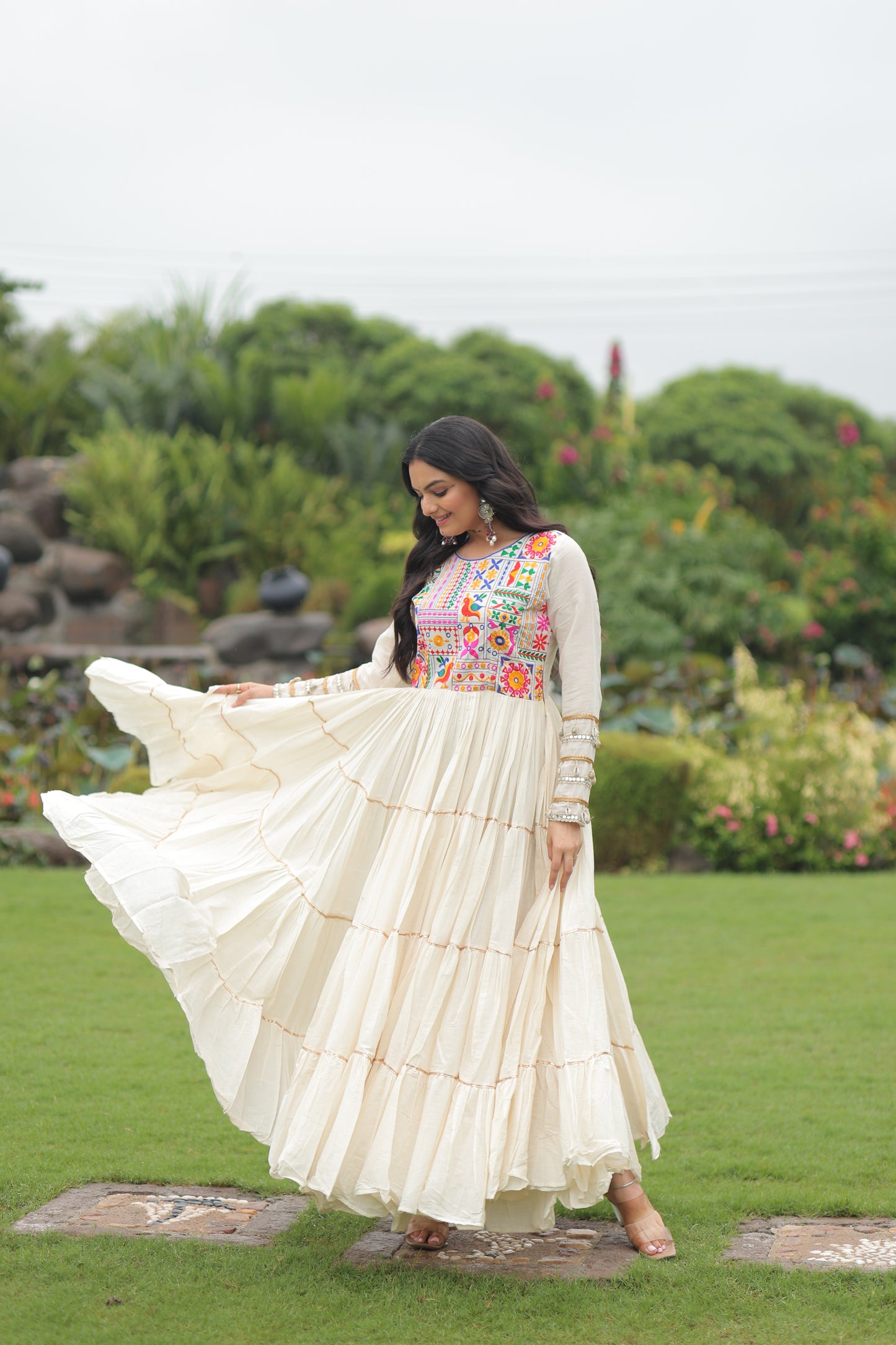 Woman in a white traditional outfit with floral patterns standing outdoors on grass.