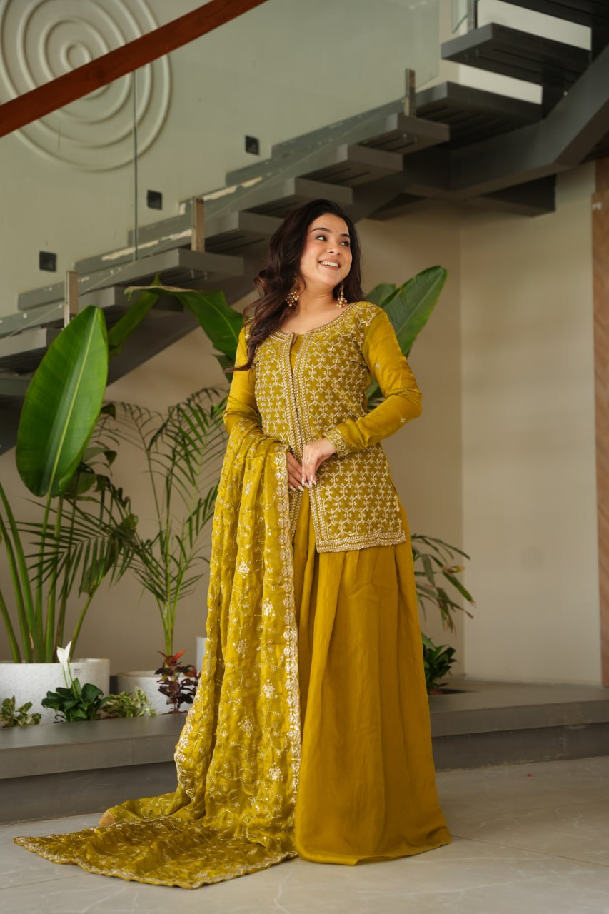 Woman in a yellow traditional outfit standing indoors with plants and a staircase in the background