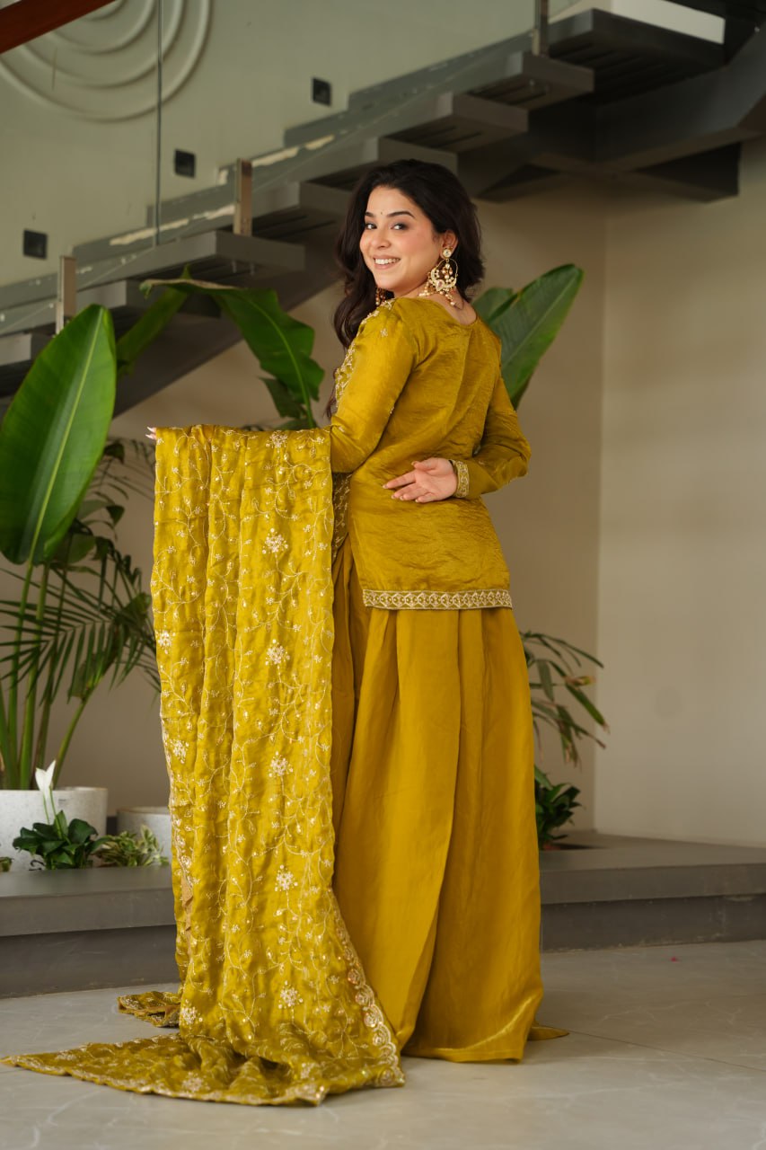 Woman in a mustard yellow traditional outfit with a matching dupatta, standing indoors.