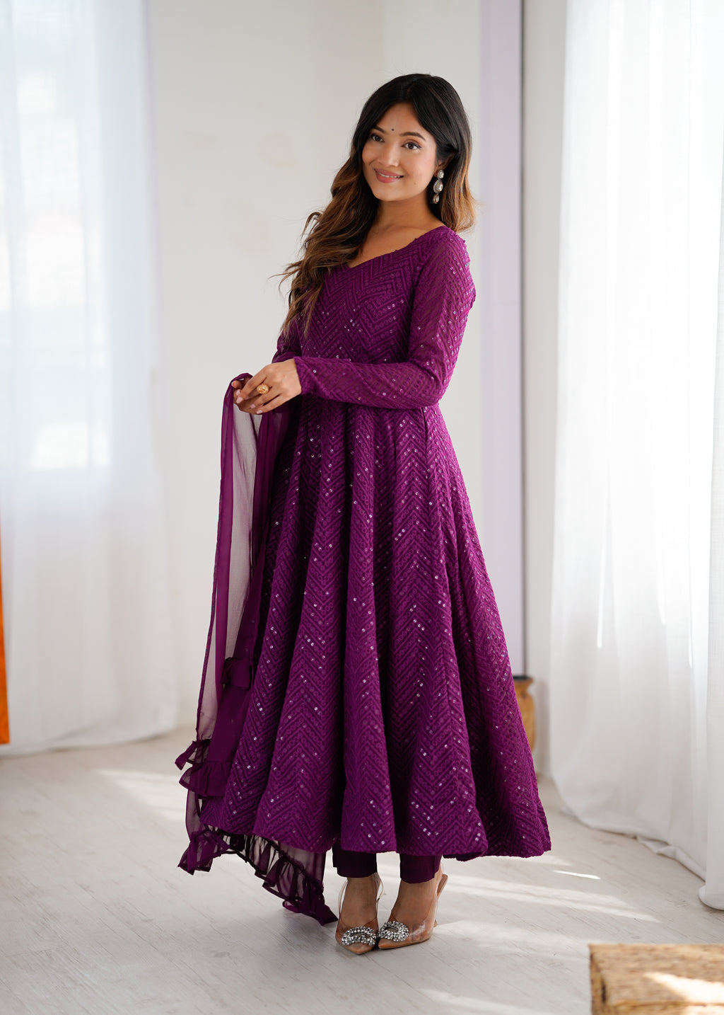 Woman wearing a purple traditional outfit with a sheer dupatta against a white curtain background