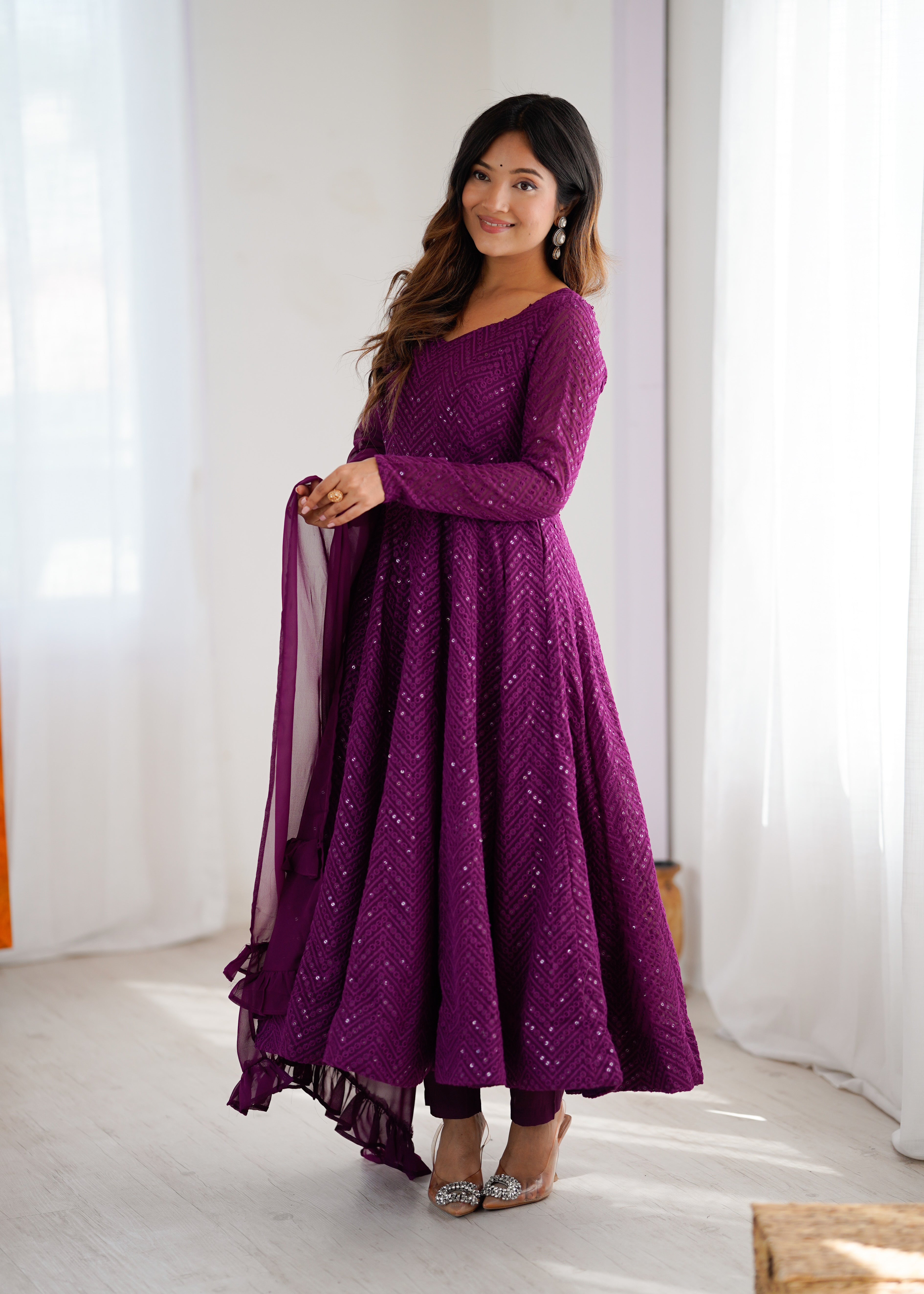 Woman wearing a purple traditional outfit with a sheer dupatta against a white curtain background