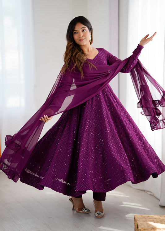 Woman in a purple dress with a matching scarf, standing indoors with white curtains in the background.