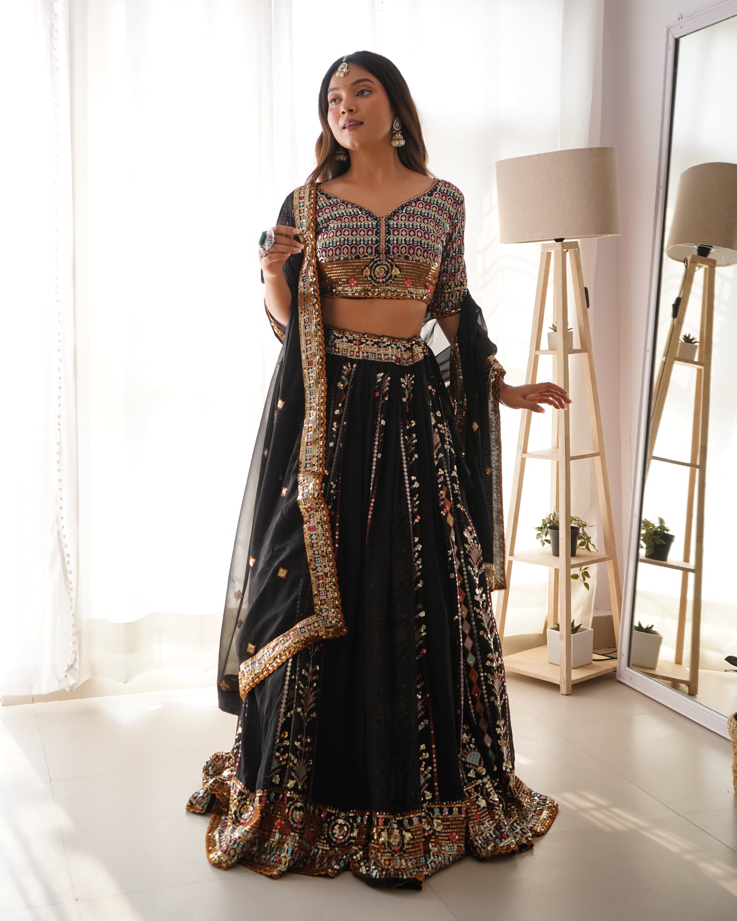 Woman in a black and gold traditional outfit standing in a bright room with a mirror and lamp.