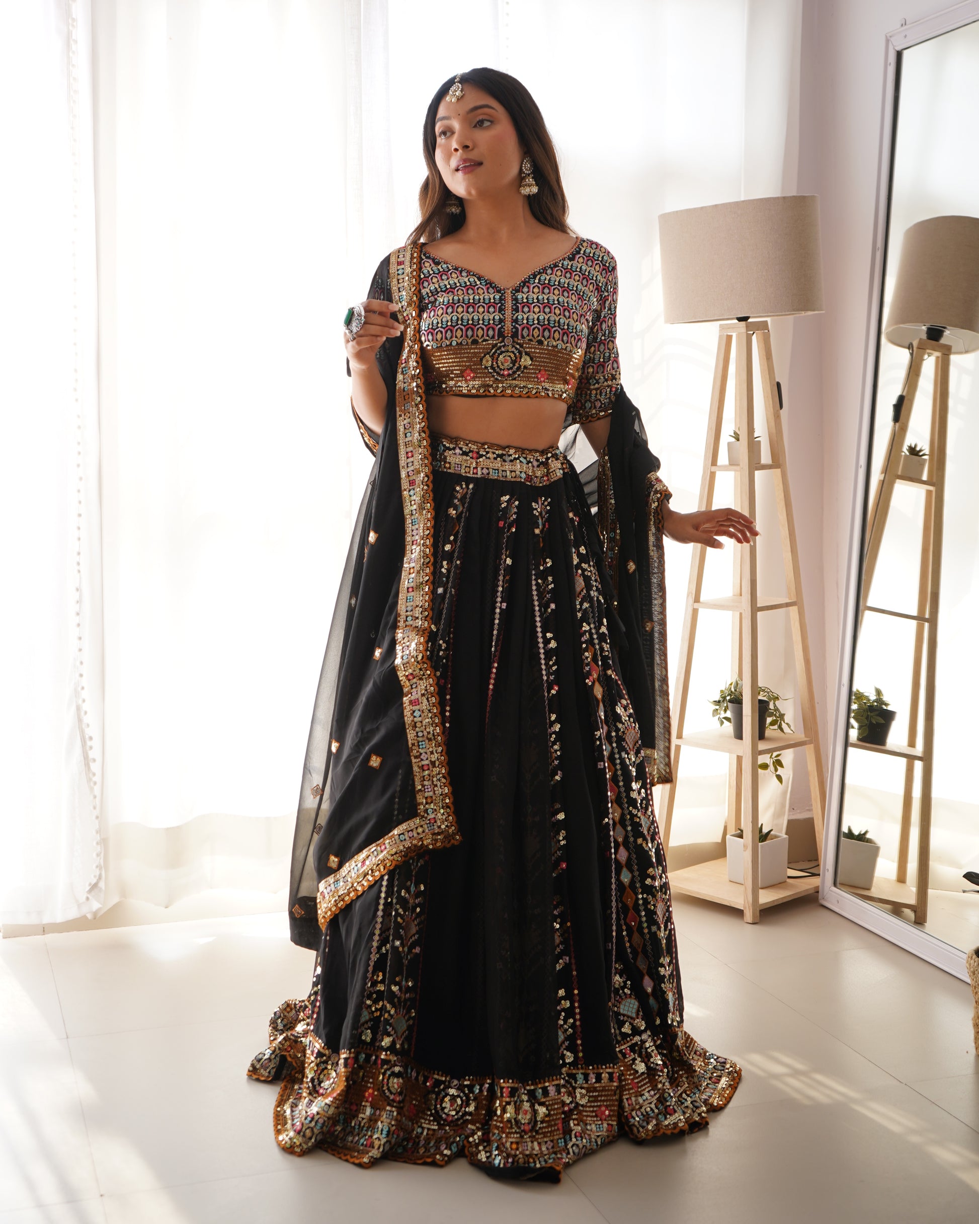 Woman in a black and gold traditional outfit standing in a bright room with a mirror and lamp.