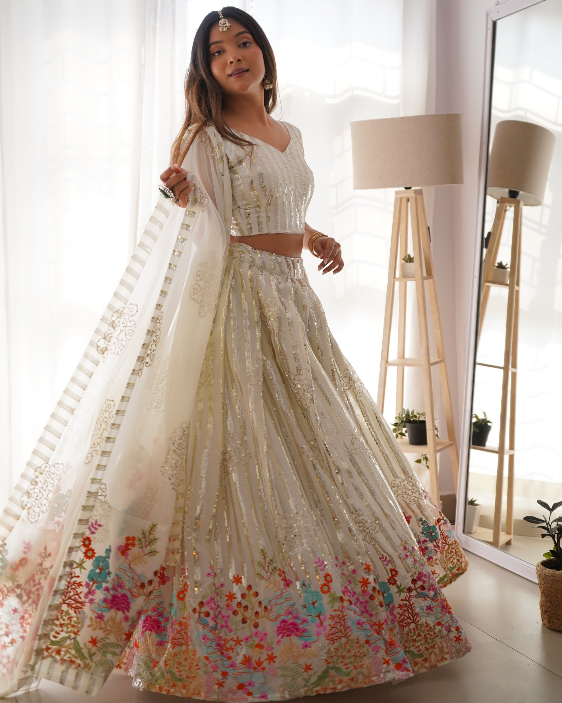 Woman in a white floral lehenga standing in a bright room with a mirror and lamp.