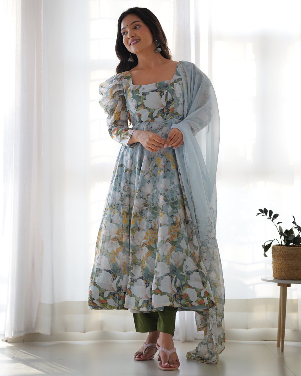 Woman wearing a floral dress with a light blue dupatta in a bright room.