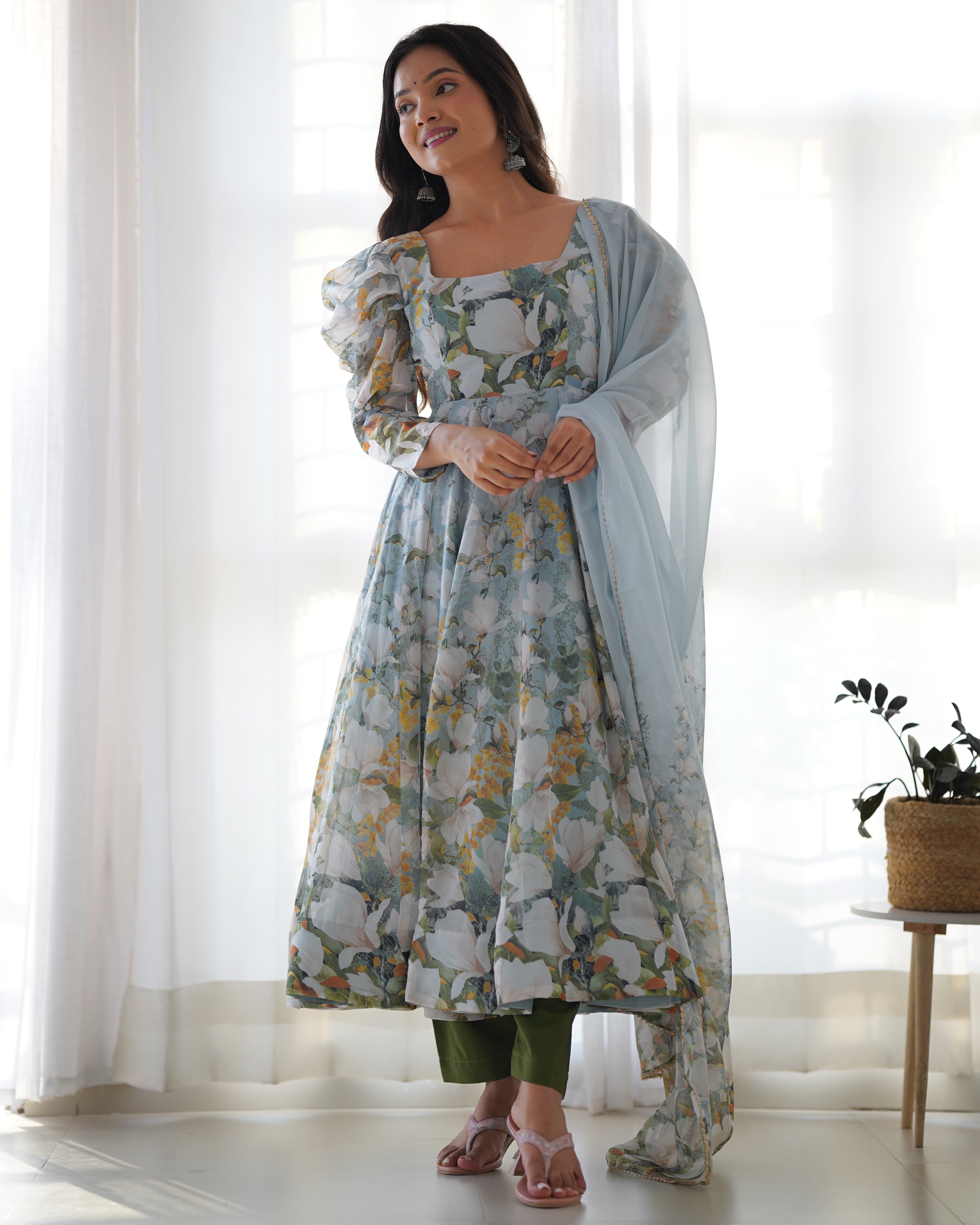 Woman wearing a floral dress with a light blue dupatta in a bright room.
