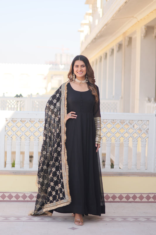 Woman in a black dress with intricate patterns standing in front of a decorative wall.