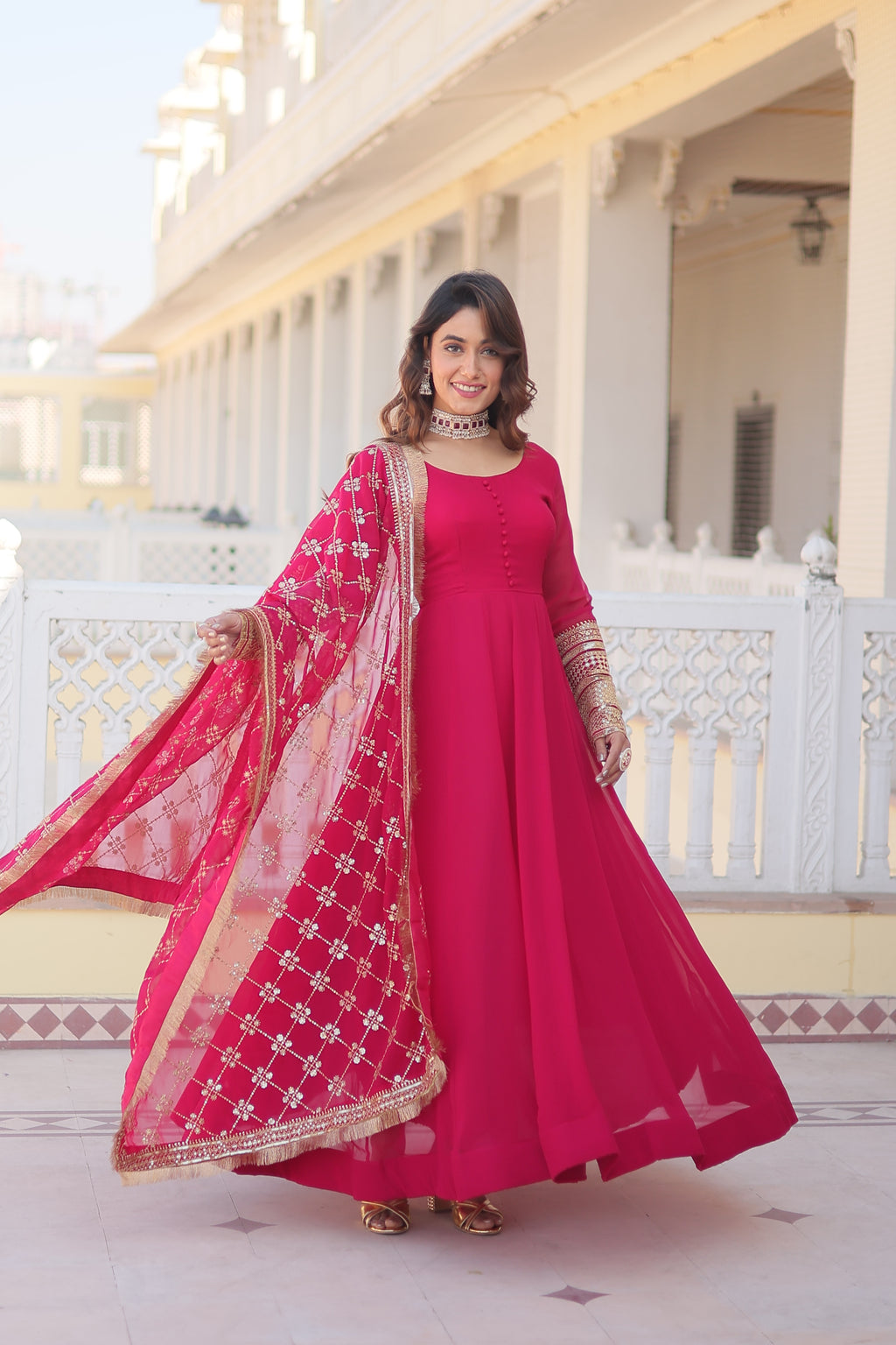 Woman in a bright pink traditional outfit with a matching dupatta standing in front of a white staircase.