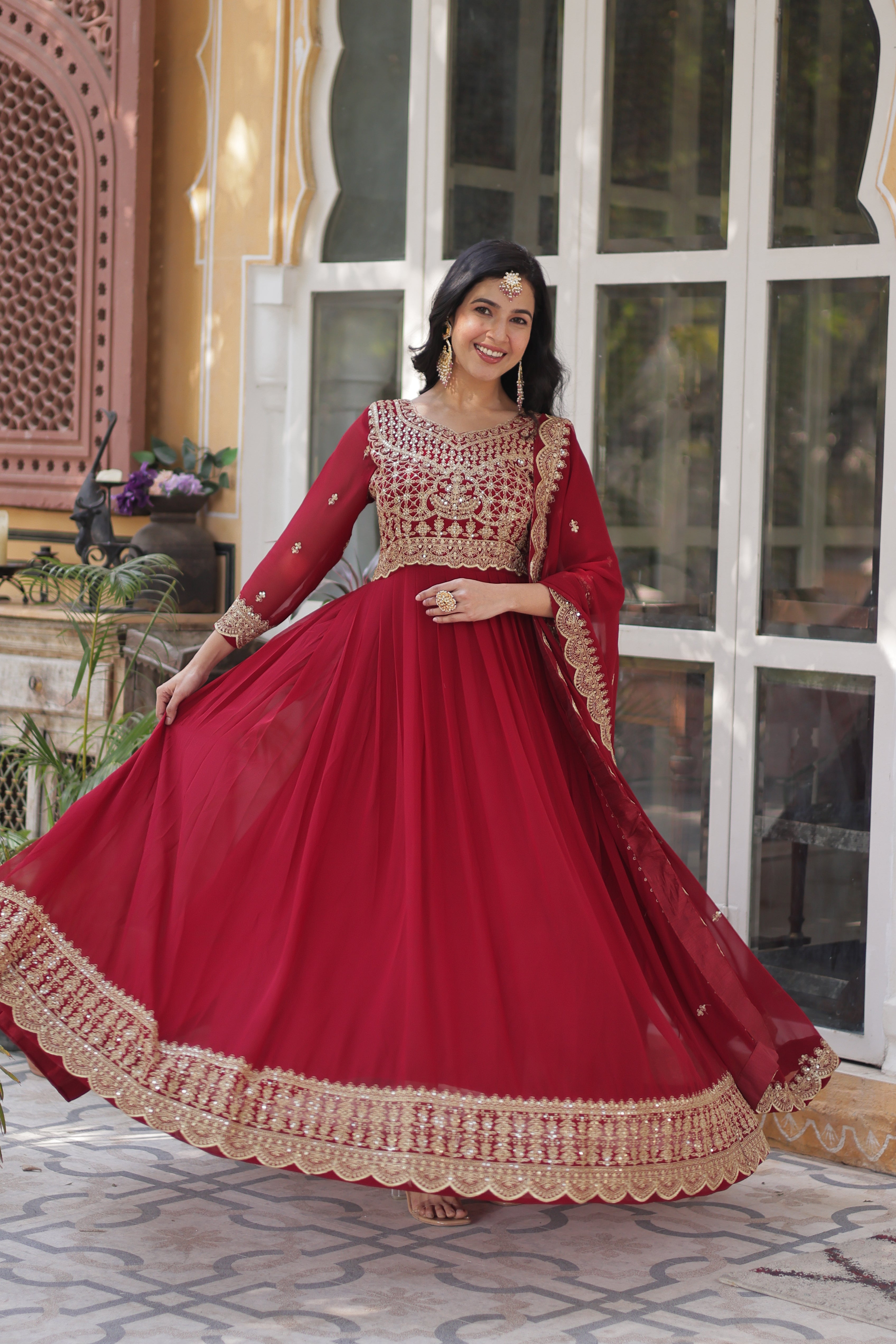 Woman in a red traditional outfit with gold embroidery standing in front of a window.