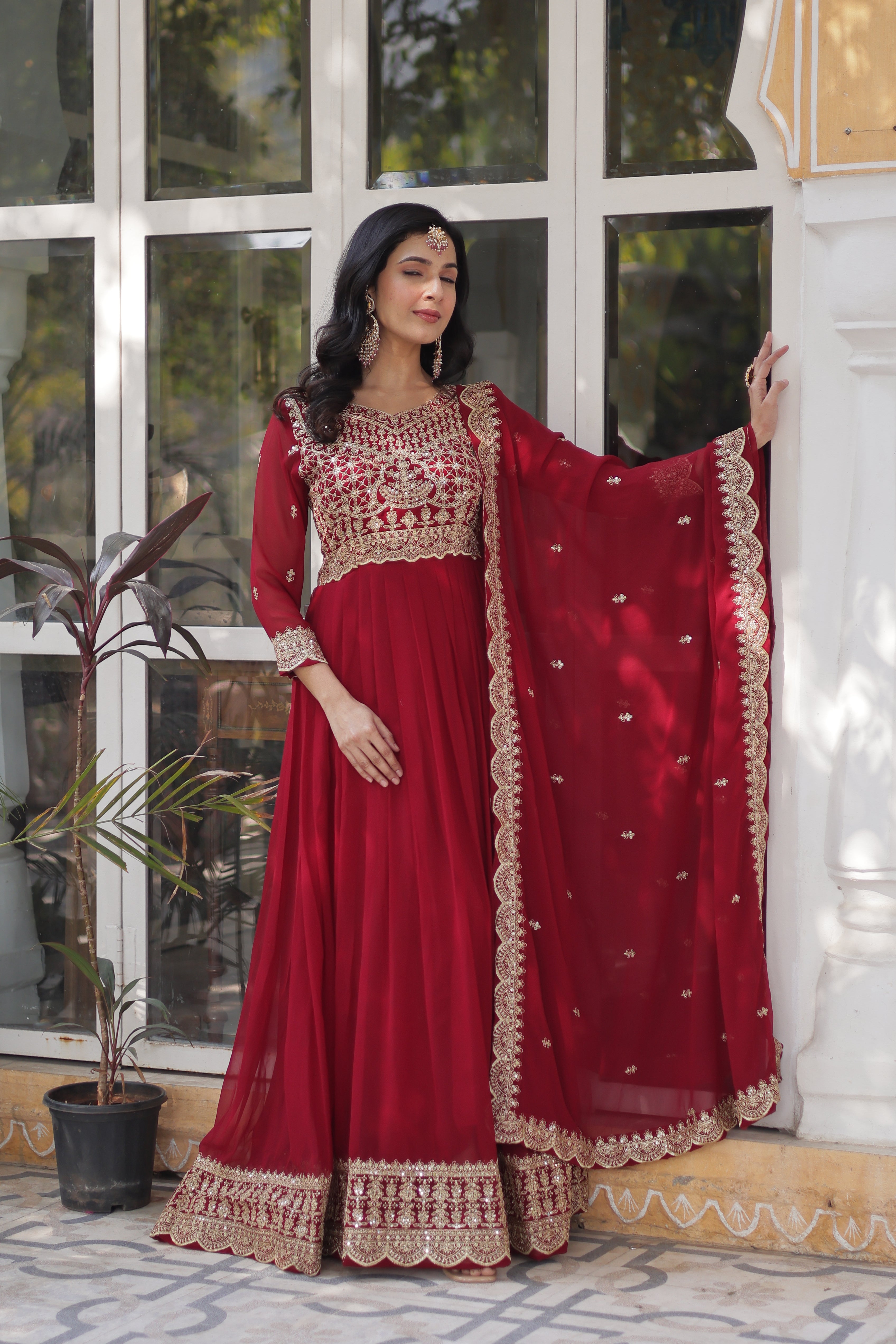 Woman wearing a red traditional outfit with intricate designs in a home setting.