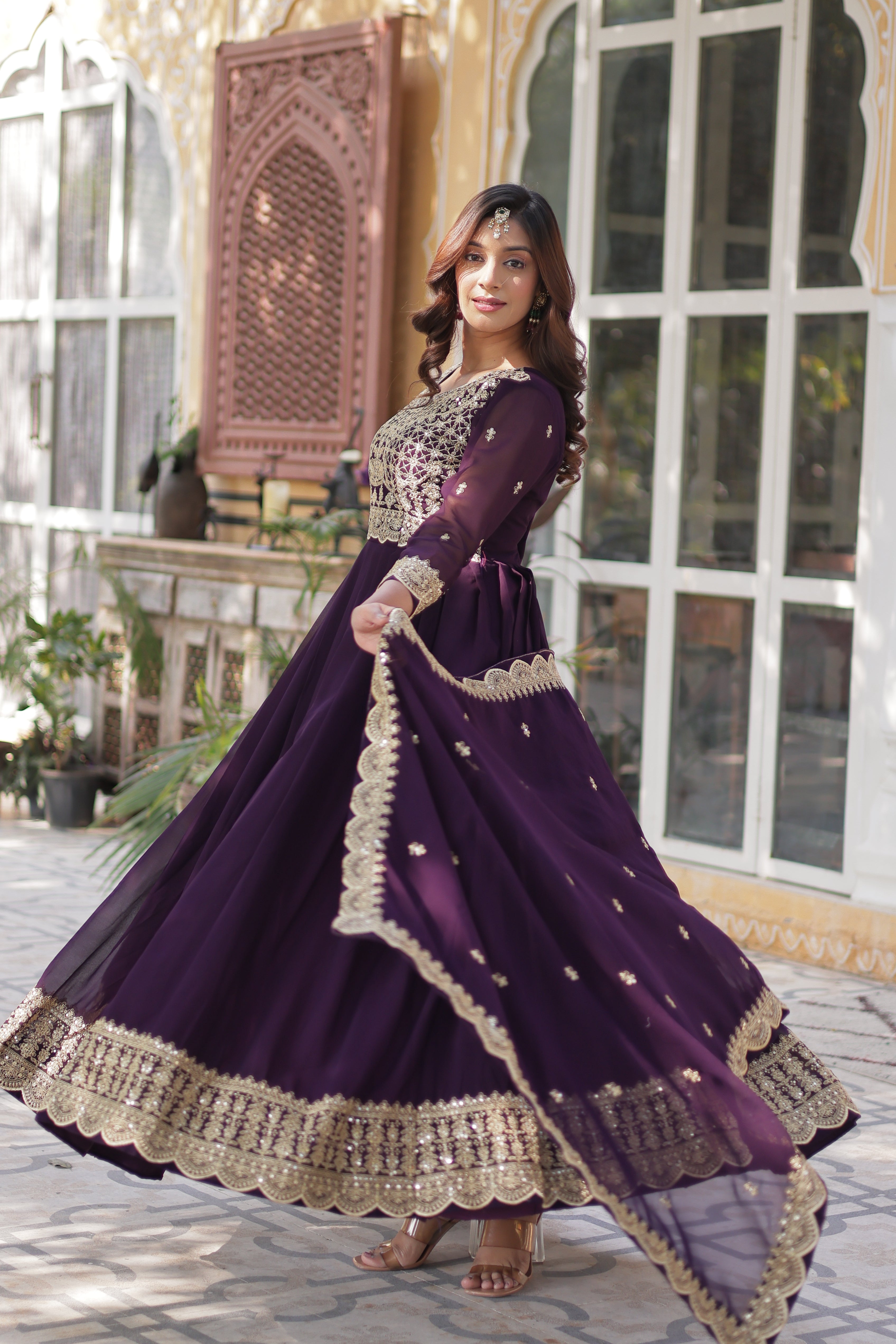 Woman in a purple traditional outfit with gold trim standing in front of a window.