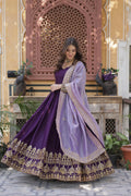 Woman in a purple traditional outfit with gold embroidery standing in front of a decorative wall.