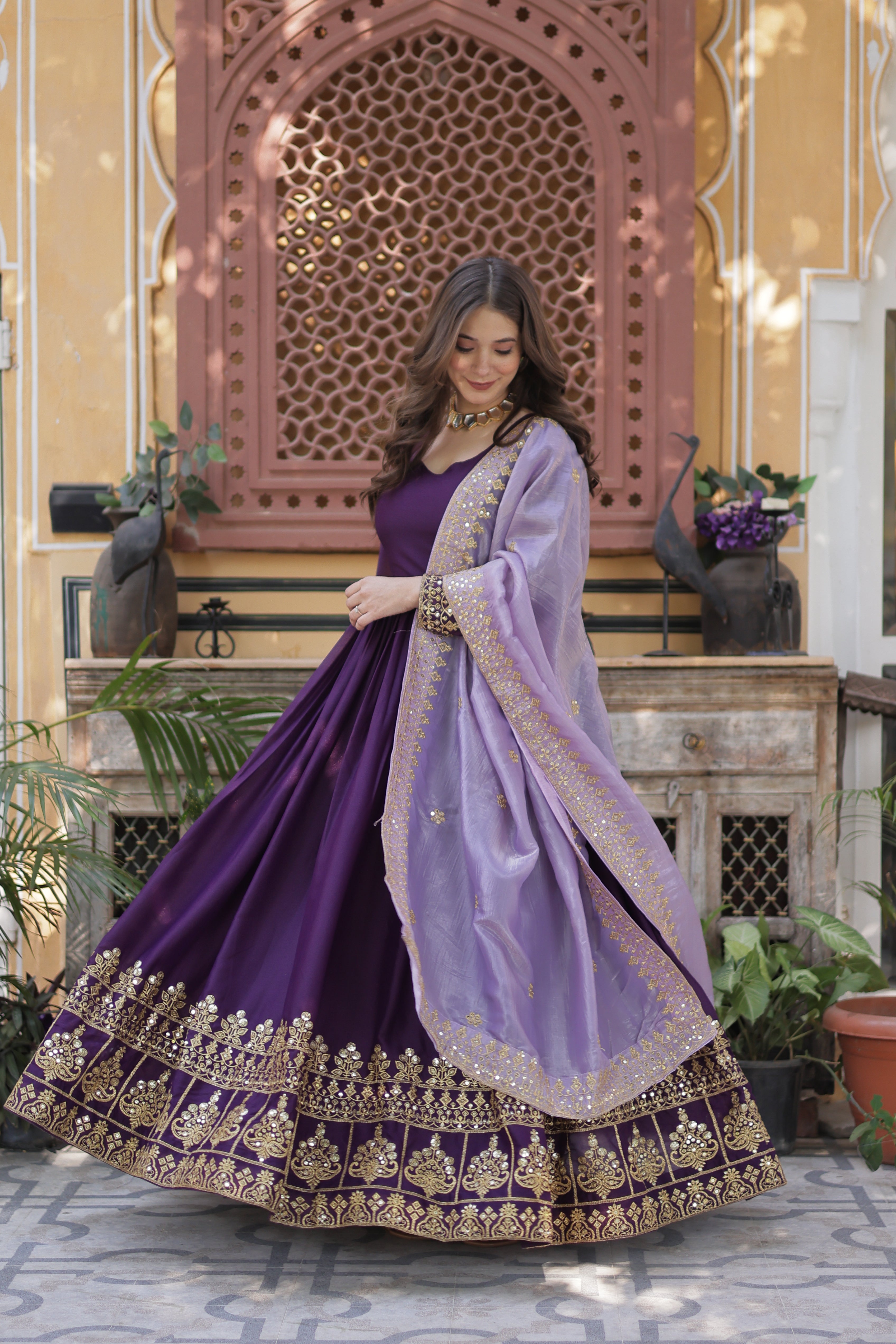 Woman in a purple traditional outfit with gold embroidery standing in front of a decorative wall.