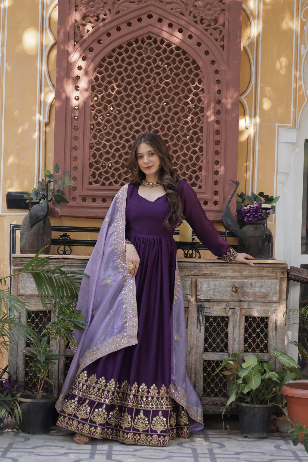 Woman in a purple dress with gold embroidery standing in front of a decorative wall.