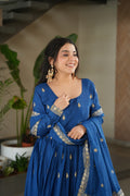 Woman in a blue traditional outfit with gold embroidery, smiling indoors.