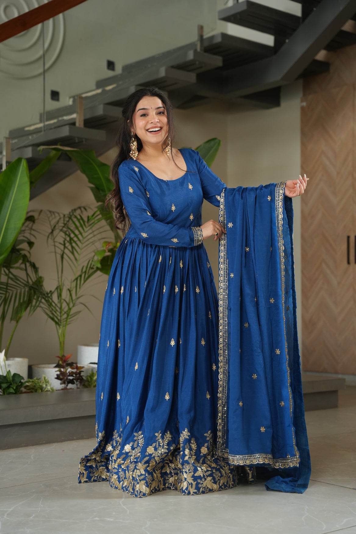 Woman in a blue traditional outfit with gold embroidery standing indoors.