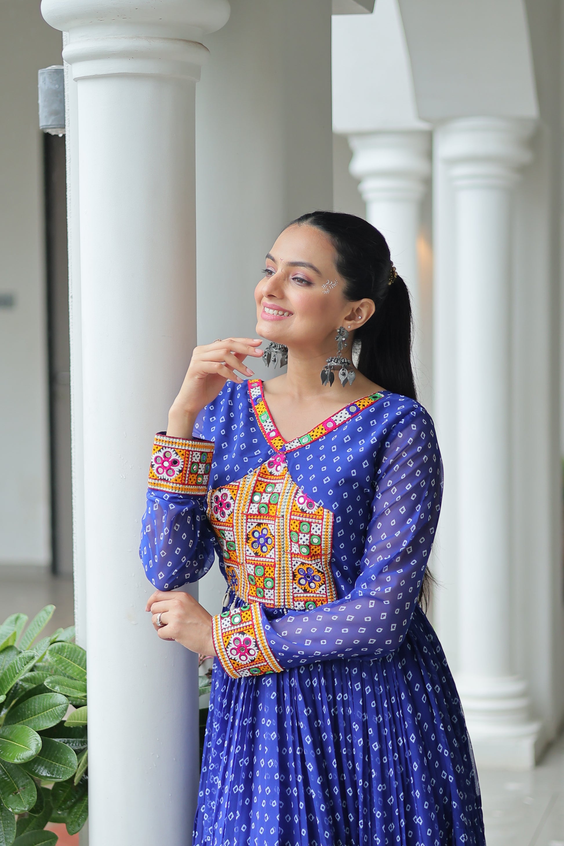 Woman in traditional blue outfit with colorful patterns leaning against a white column.