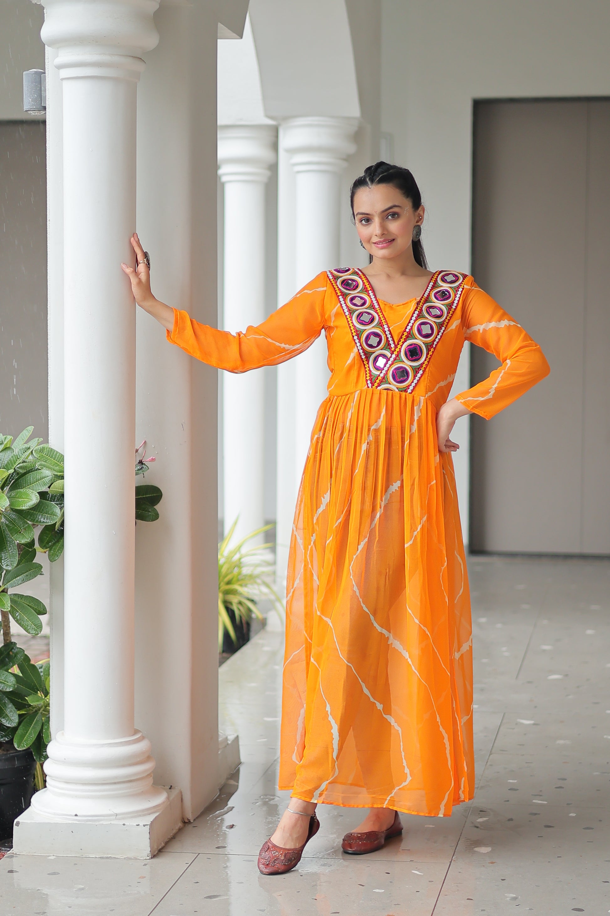 Woman in an orange traditional outfit standing indoors with white columns and greenery.