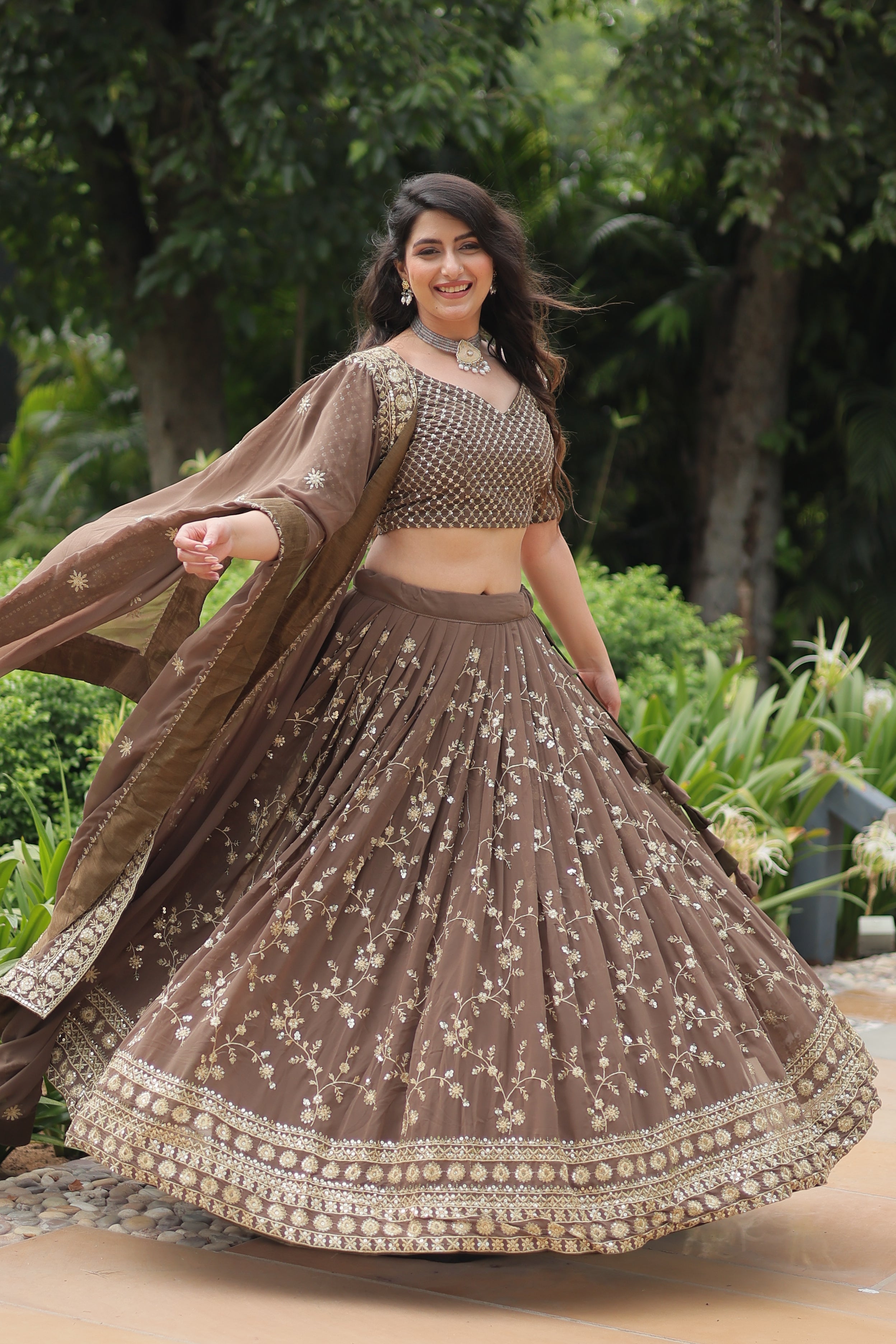 Woman in a brown traditional outfit with intricate patterns standing outdoors with greenery in the background.