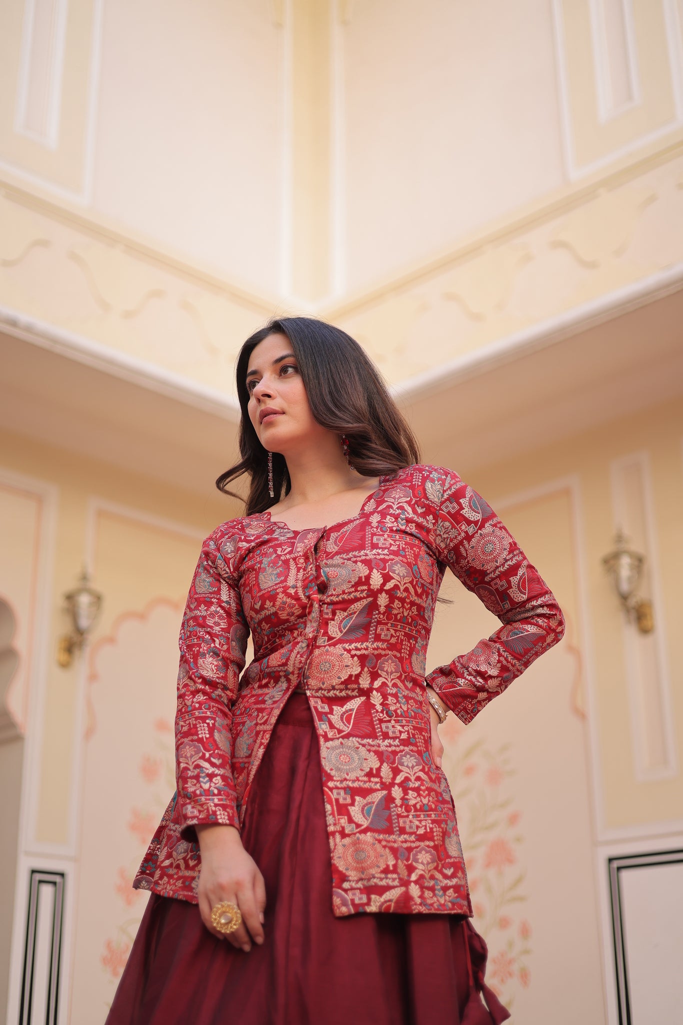 Woman wearing a red and gold traditional outfit in a room with decorative walls.
