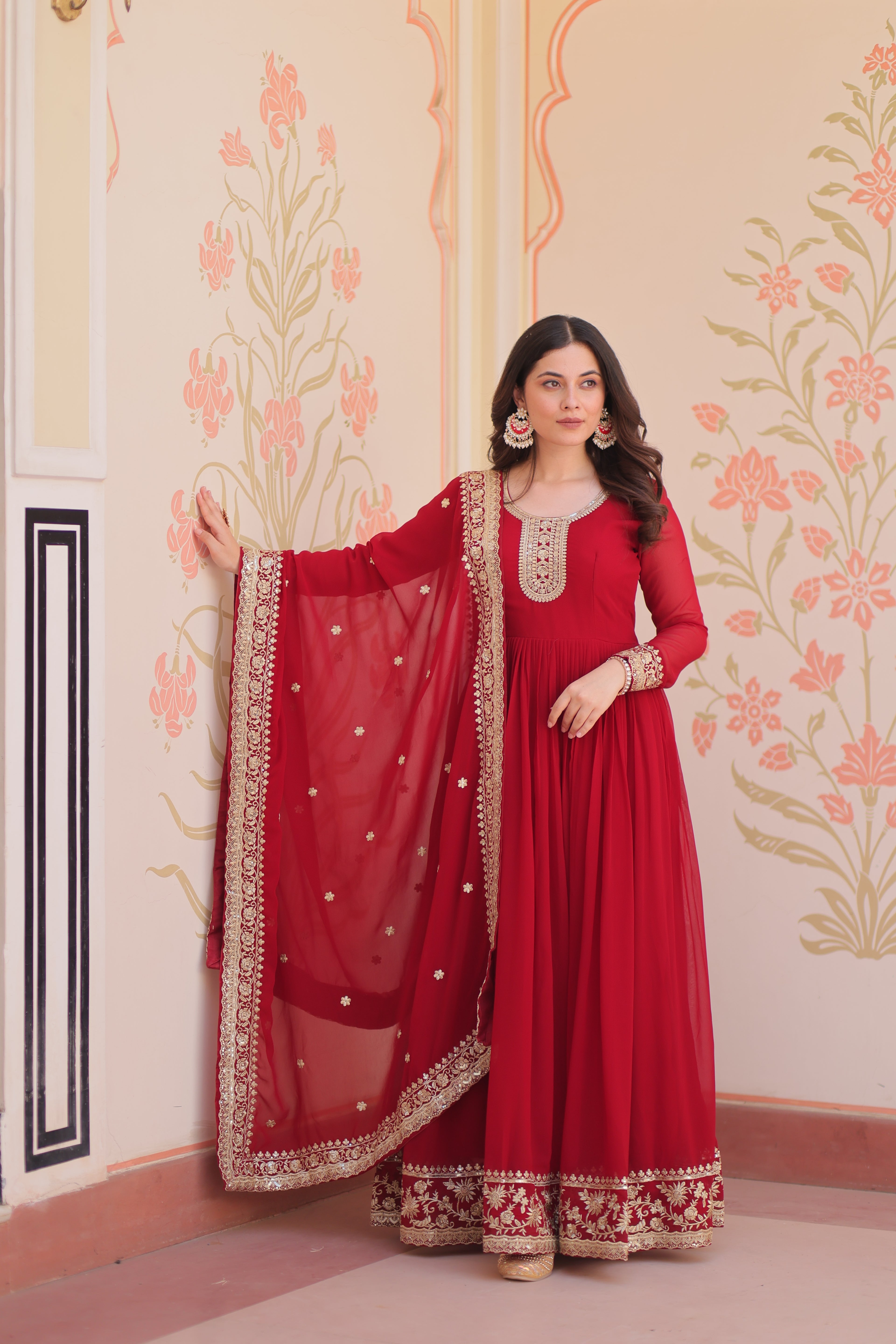 Woman in a red traditional outfit with gold embroidery standing against a decorative wall.